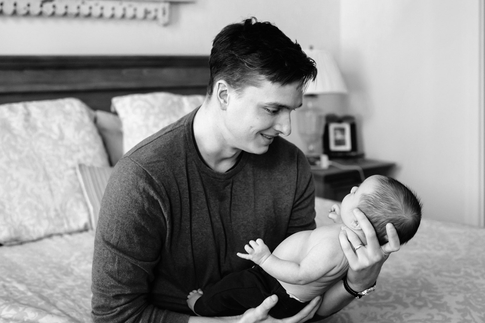 A black and white image of a new father sitting on his bed and smiling down at his newborn son in his arms during a home newborn photo session