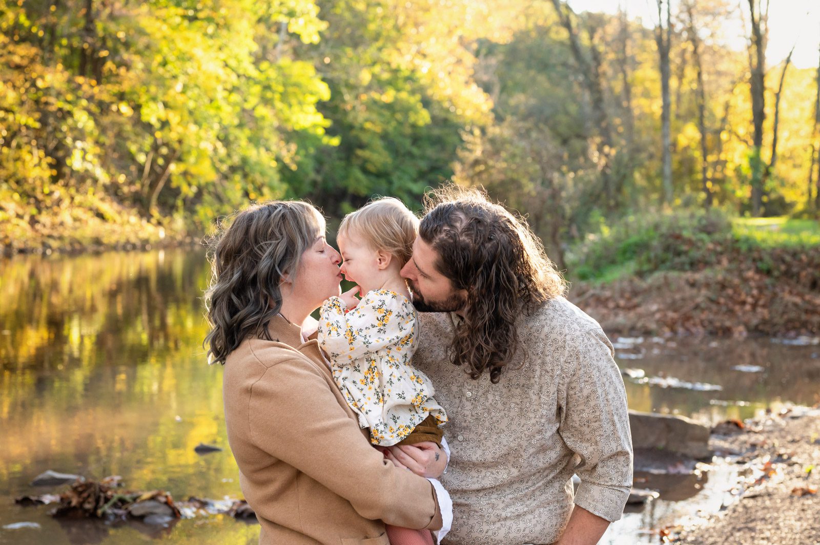 Parents standing on the edge of a creek and kissing their young daughter during a family photoshoot