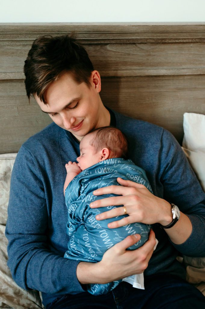 A father snuggling his newborn son against his chest and smiling down at him during a home newborn photo session