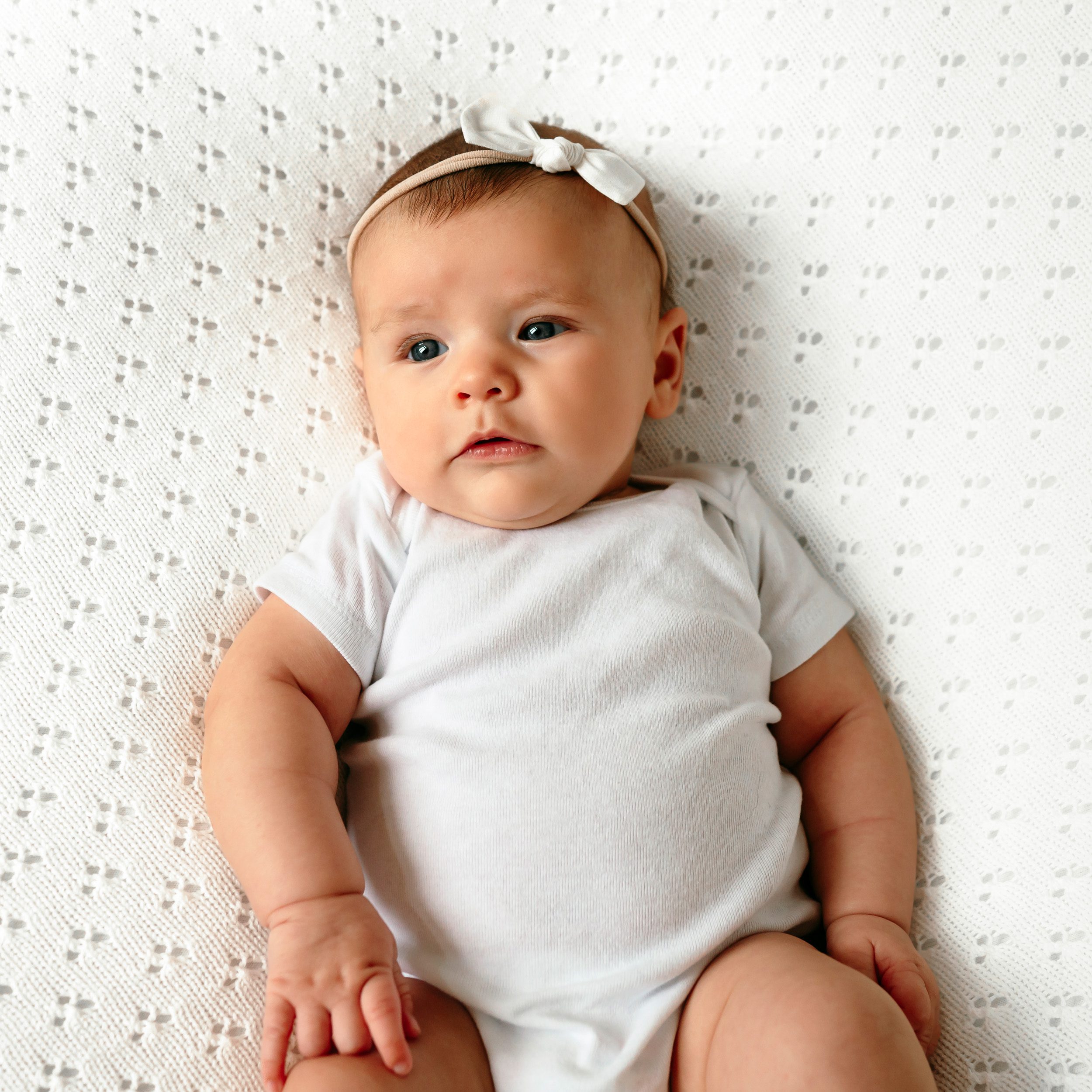 A baby girl in a white onesie on a white textured backdrop looking toward the window light at a home newborn photoshoot