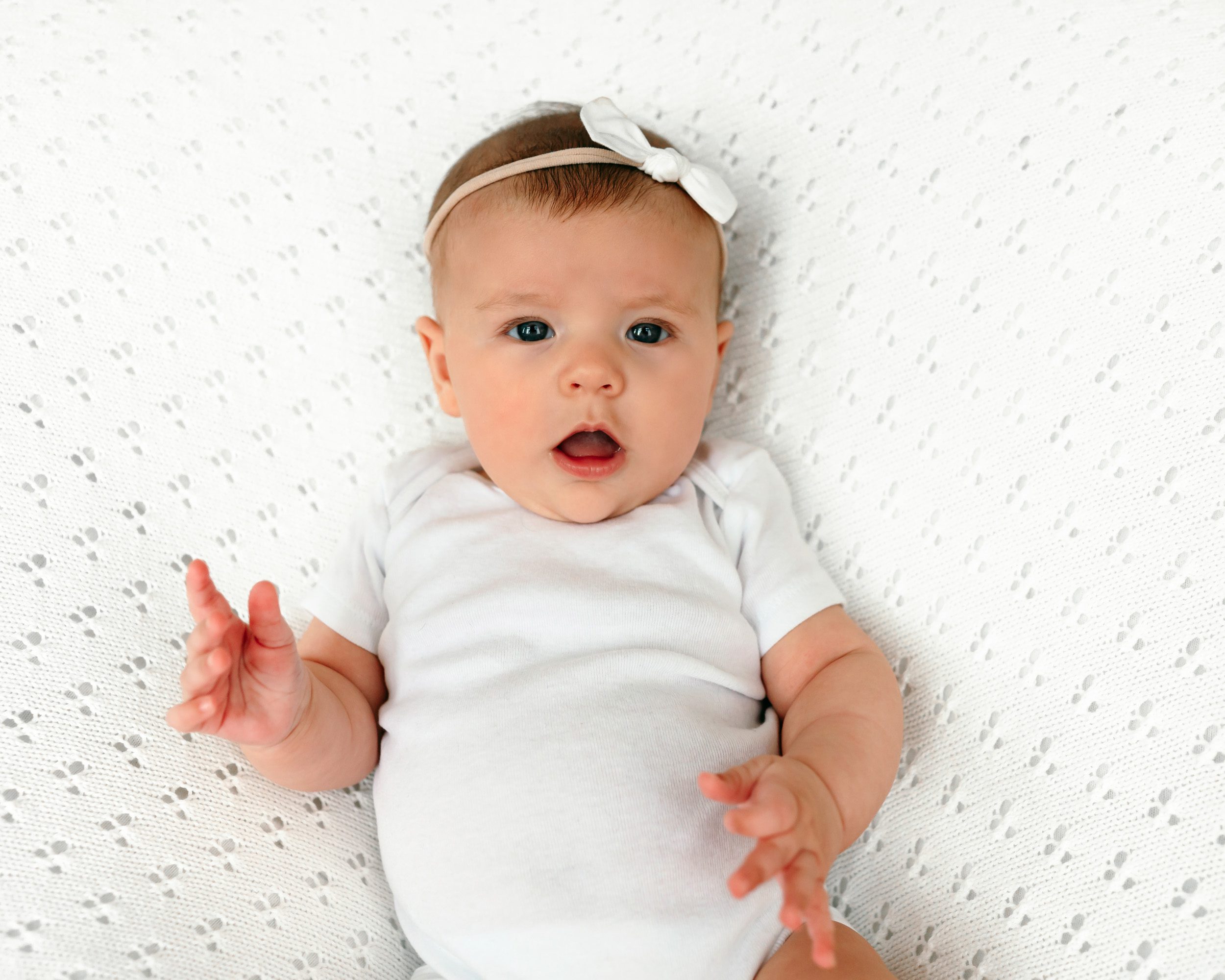 A baby girl in a white onesie on a white textured backdrop looking at the camera with a surprised expression during a home newborn photo session