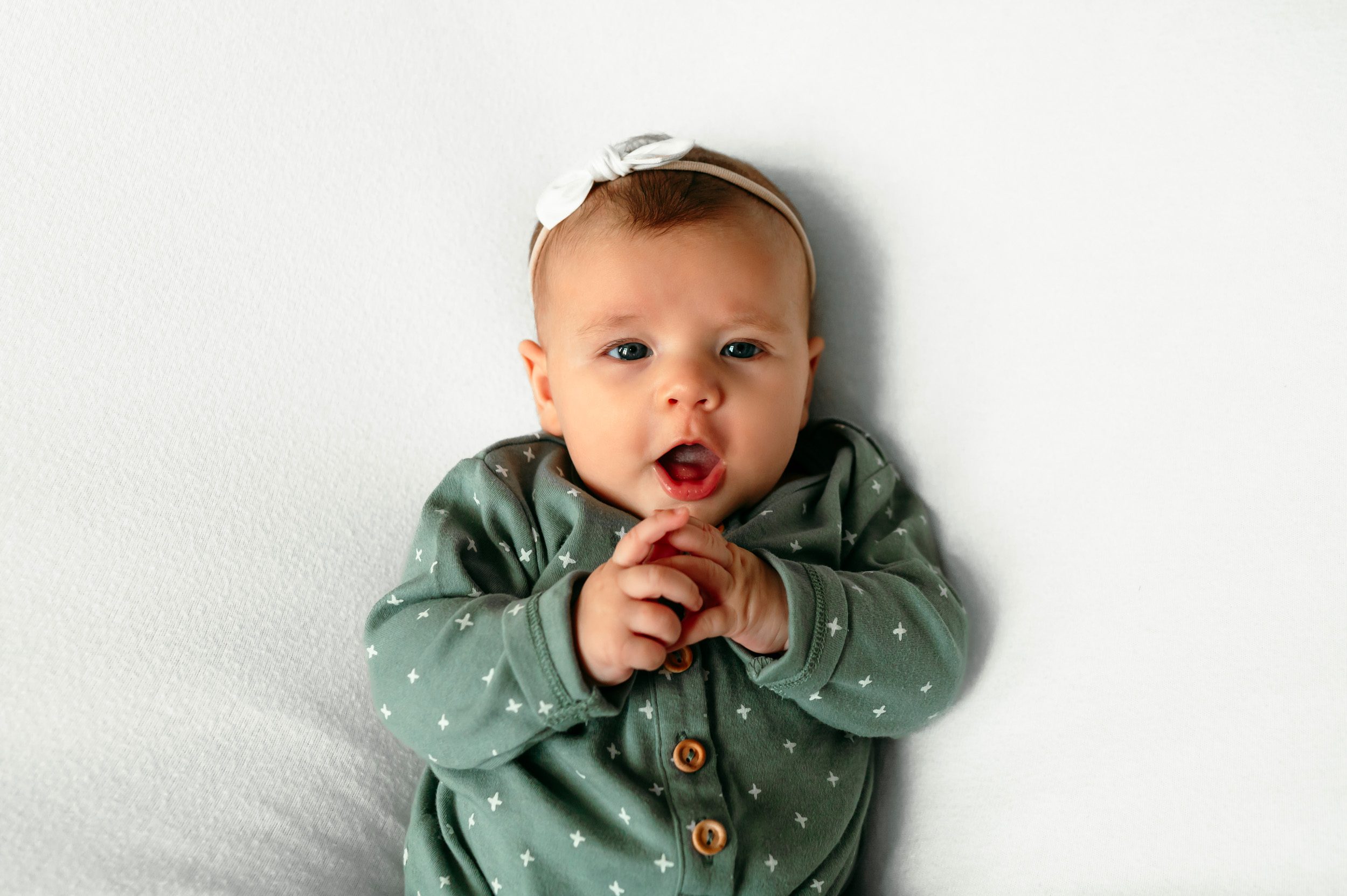 Baby girl in a green sleeper on a white backdrop looking up at the camera with a surprised expression at a home newborn photoshoot