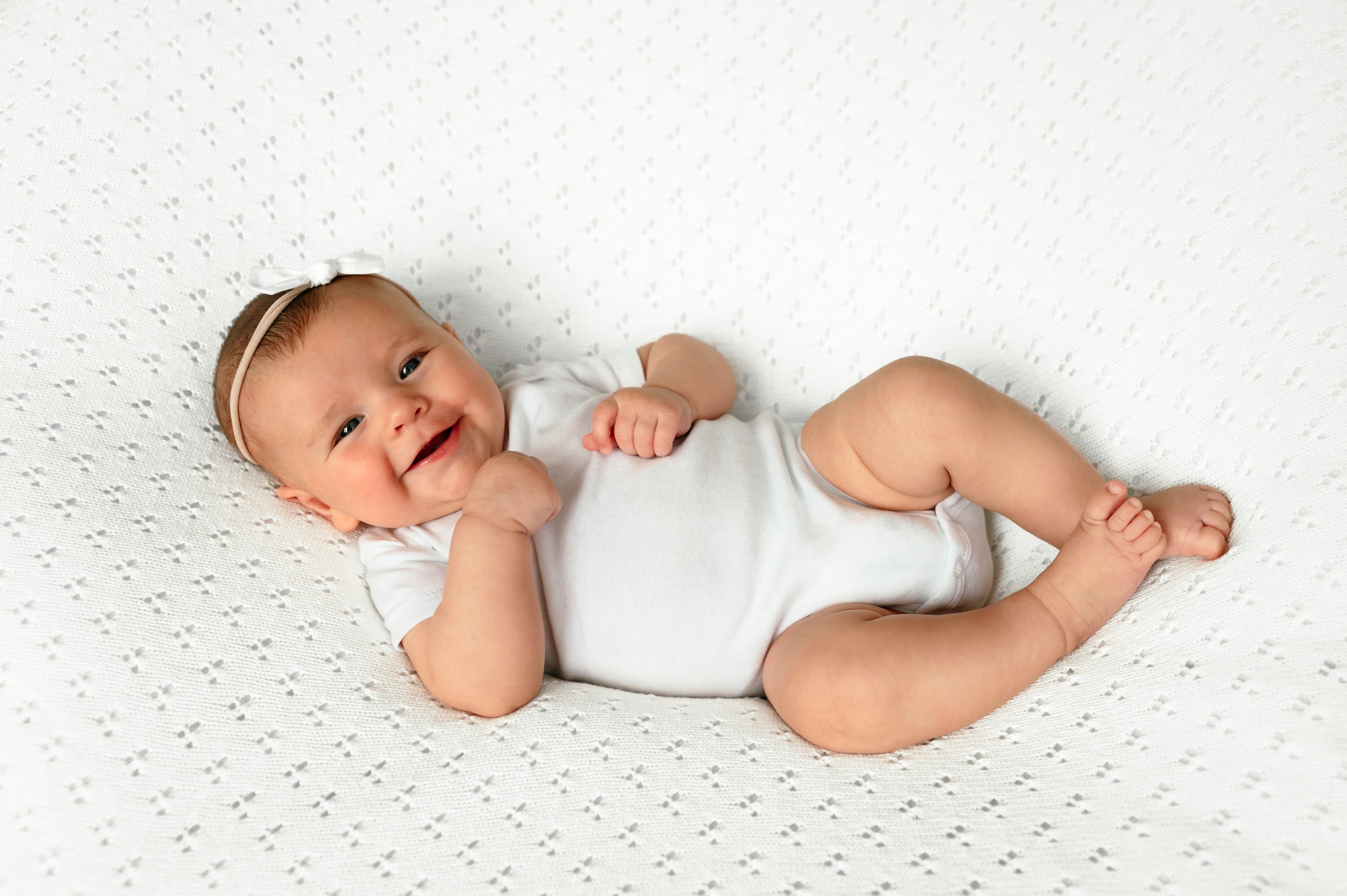 Baby girl laying on a white textured backdrop and smiling up at the camera at a home newborn photoshoot