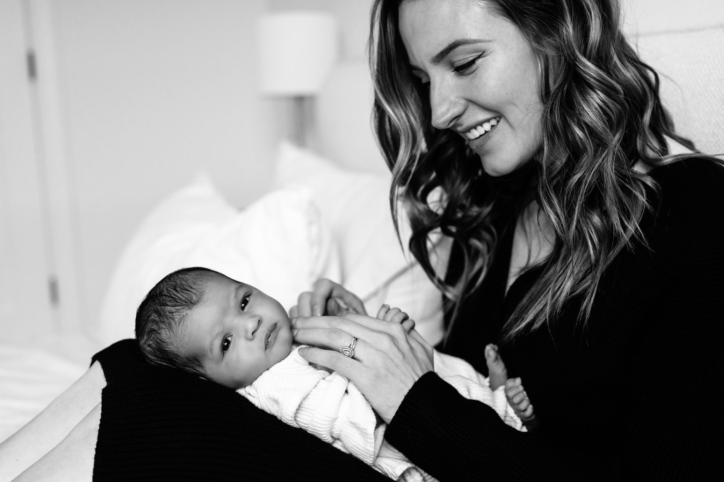 black and white image of newborn baby girl laying on her mom's legs and looking at the camera while her mom smiles down at her at an in home newborn photoshoot