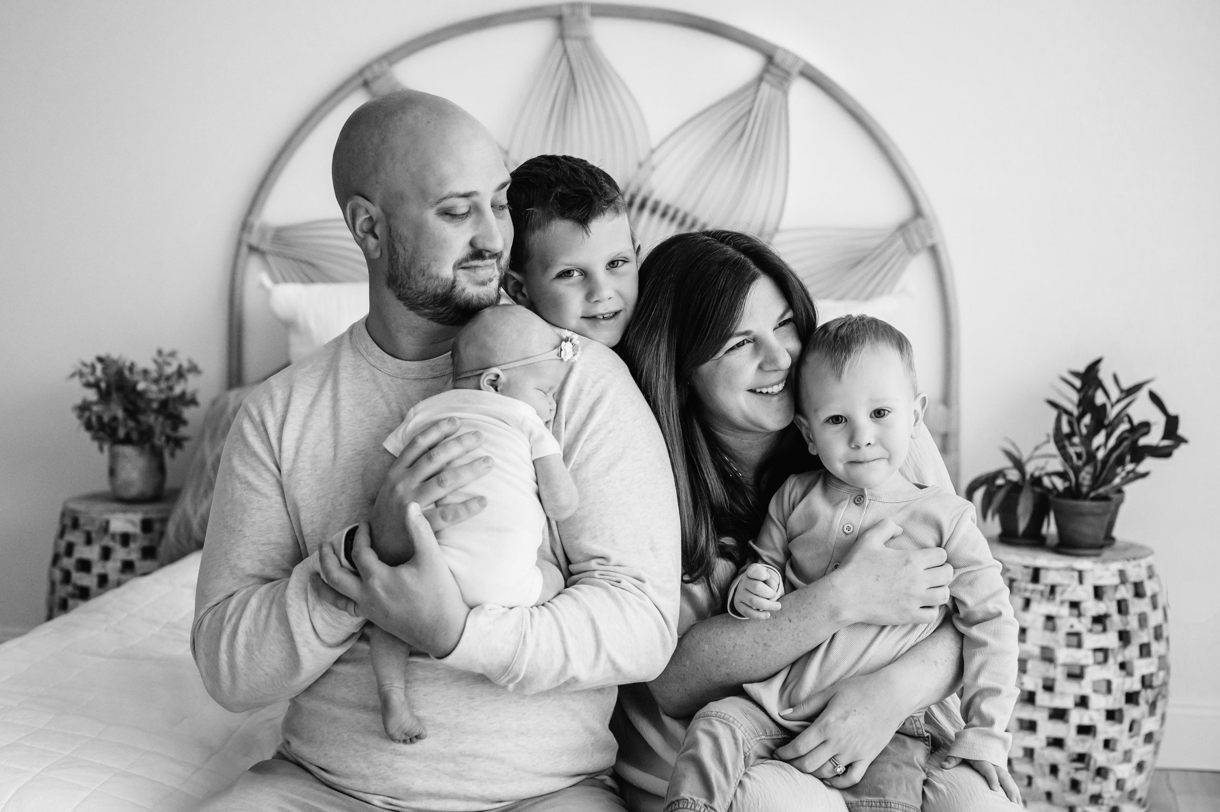 a black and white picture of parent sitting at the foot of a bed while dad holds his newborn daughter against his chest and the two older sons snuggle with mom during a lifestyle newborn photography session