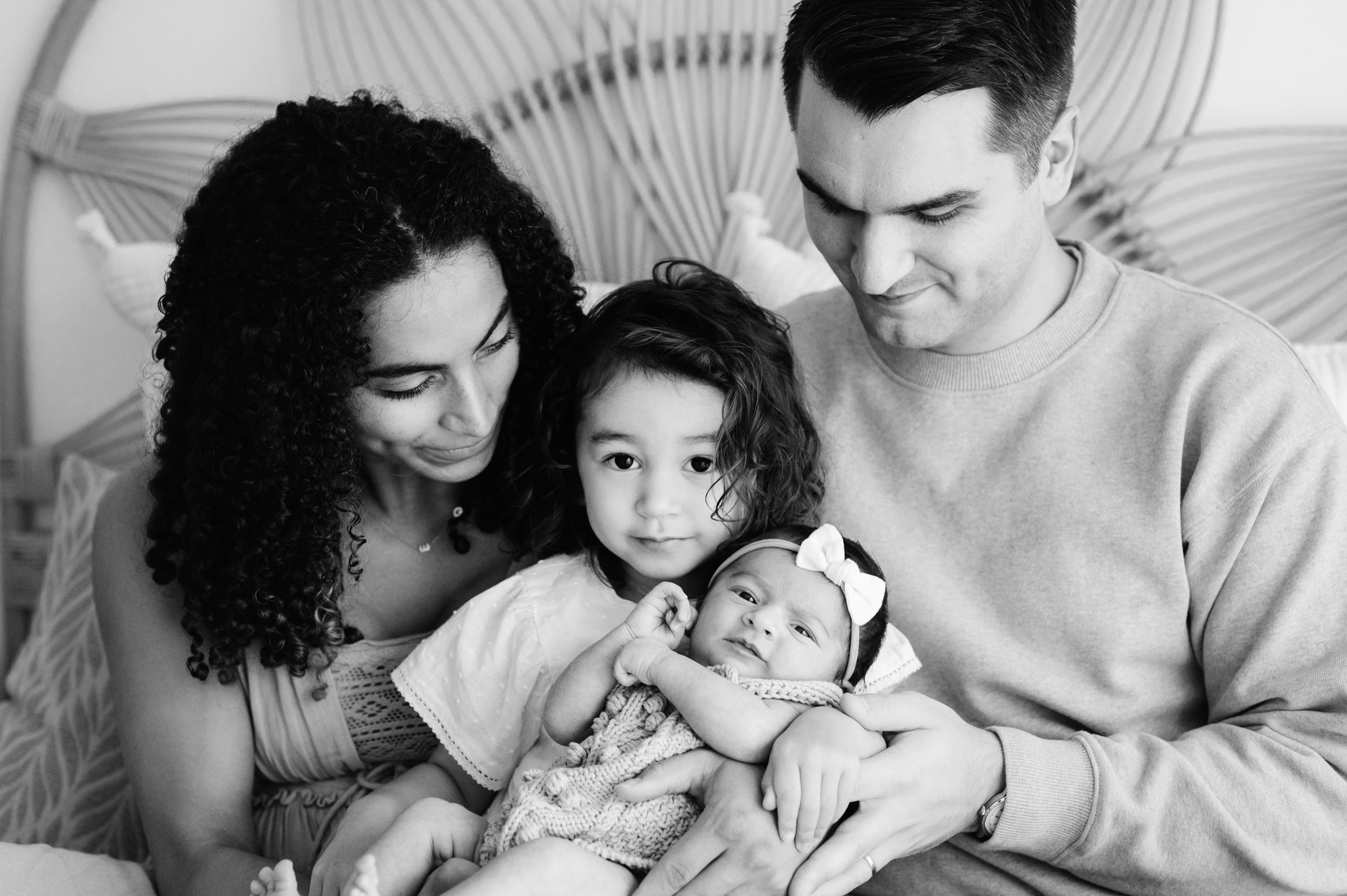 a black and white picture of young girl holding her baby sister in her arms as she sits on her mom and dad's lap during a lifestyle newborn photography session
