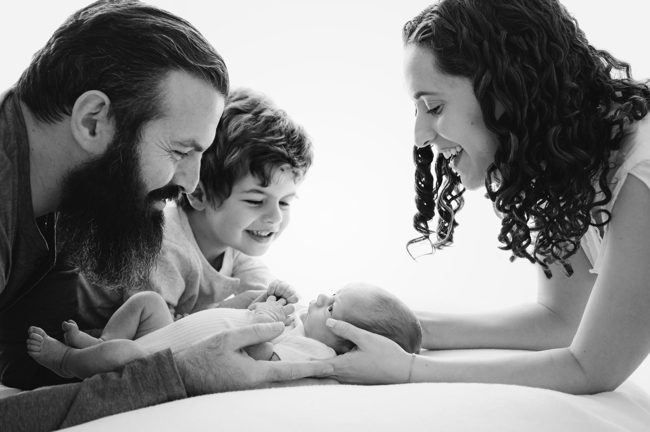 a black and white backlit photo of a baby boy laying on a bean bag with his parents and big brother smiling down at him during a lifestyle newborn photoshoot