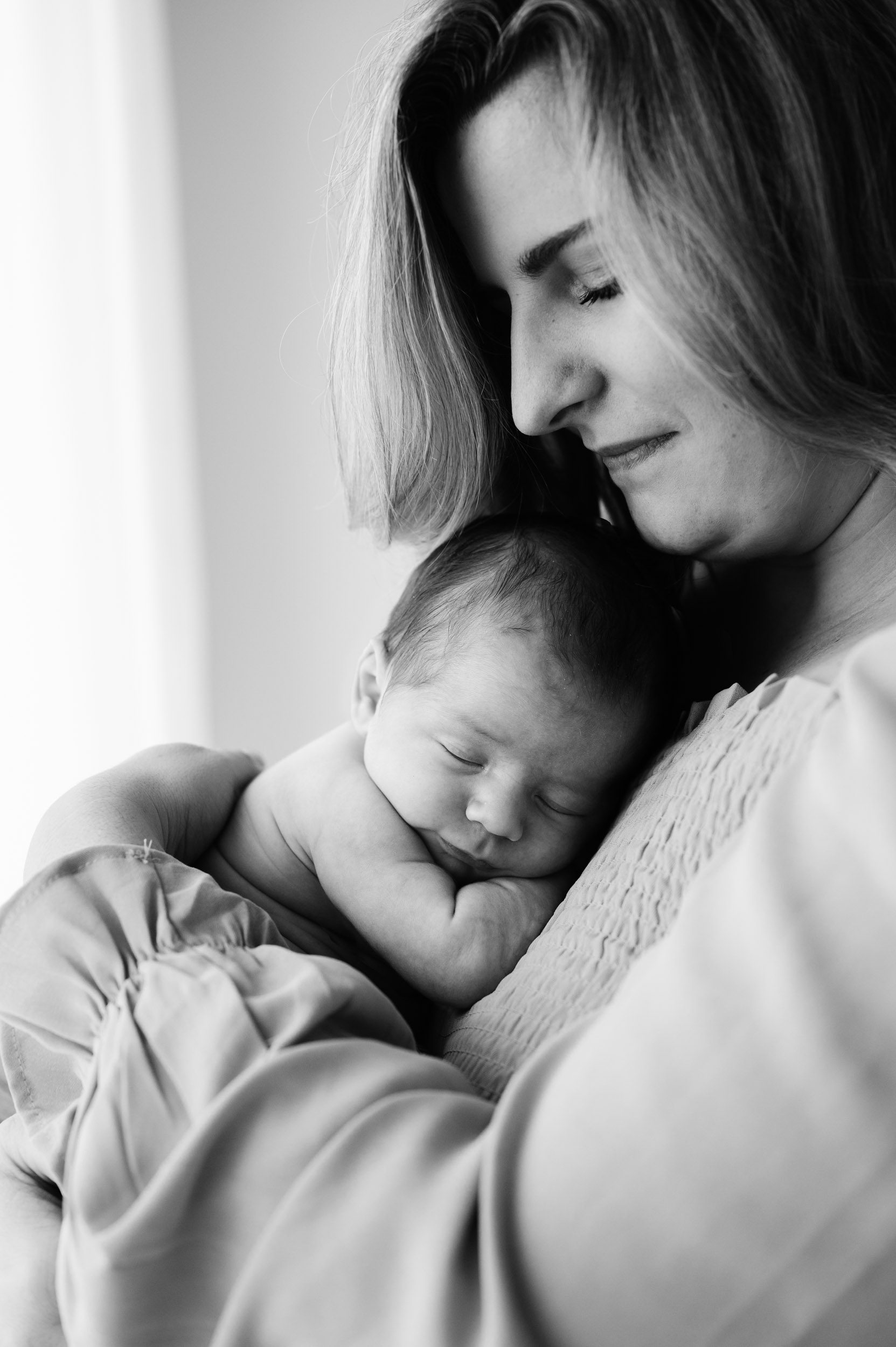 a black and white picture of a new mom holding her baby boy snuggled up against her chest and smiling down at him during a natural newborn photoshoot