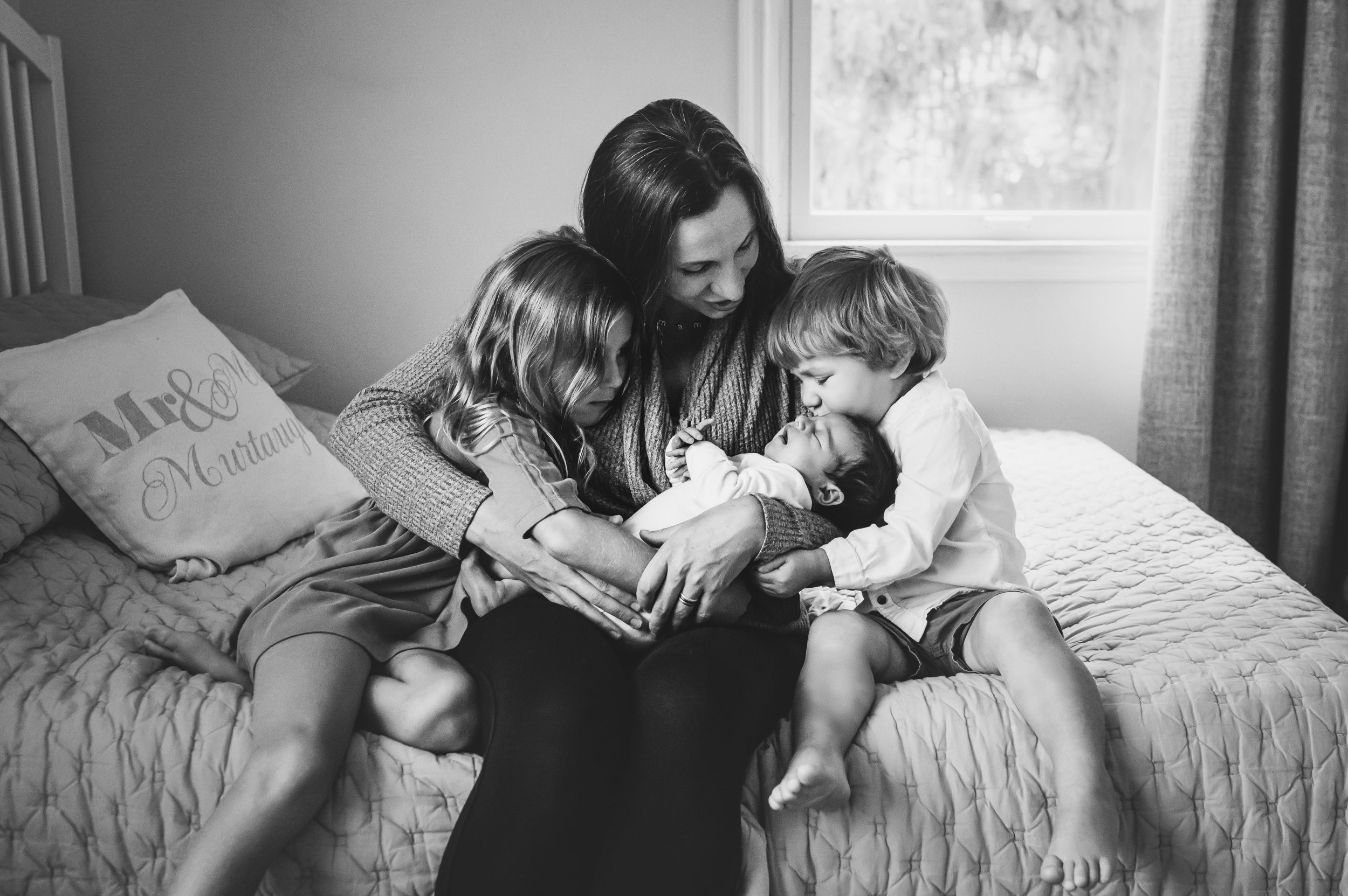 a black and white picture of a mom sitting the side of a bed cradling her newborn baby in her arms as her two older children snuggle in on both sides and her son kisses the baby on the head during a lifestyle newborn photoshoot