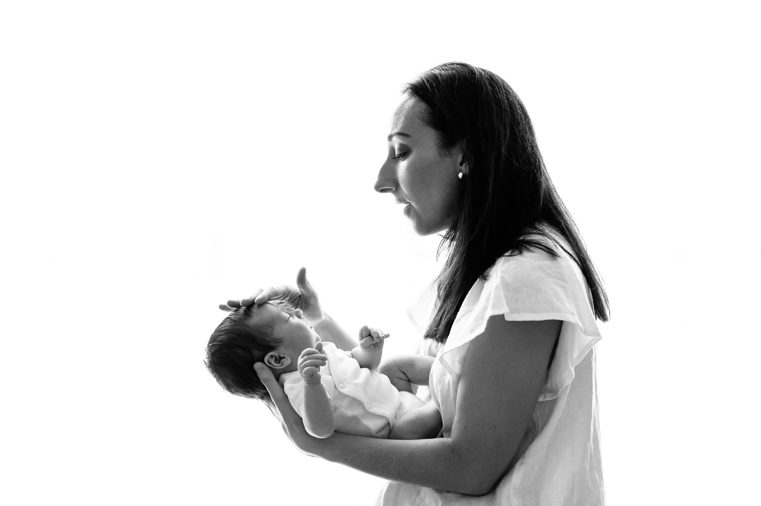 a black and white backlit picture of a new mom holding her baby boy in her arms and touching him gently on the head during a natural newborn photos session