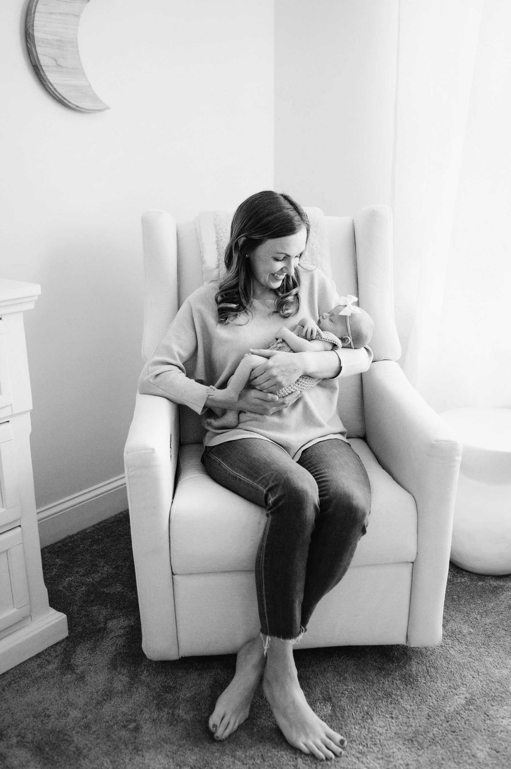 a black and white picture of a new mom sitting in a rocking chair in the baby's nursery and smiling down at her baby girl cradled in her arms during a lifestyle newborn photography session