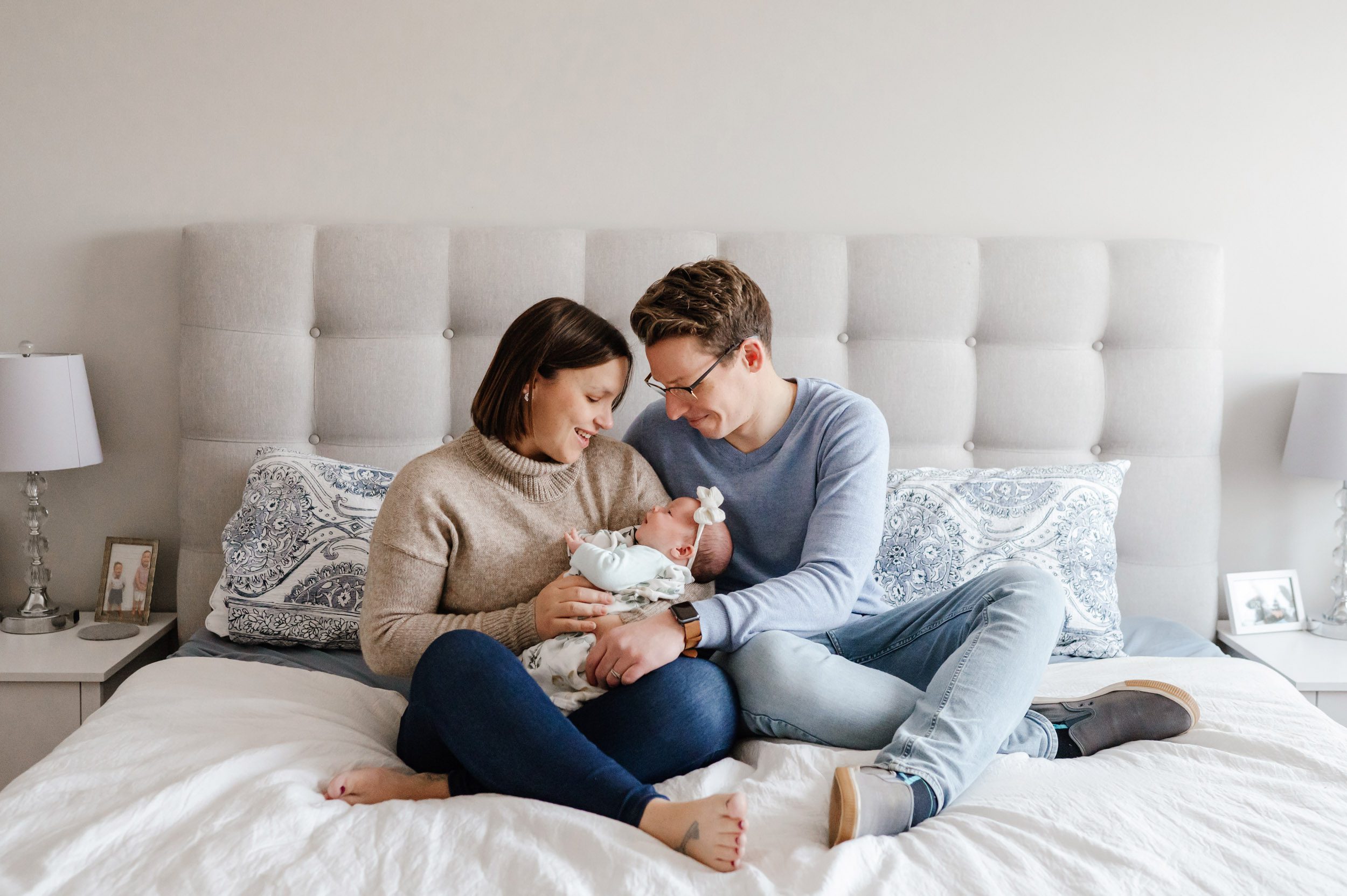 new parents sitting on a bed with their baby girl in their arms and smiling down at her during a natural newborn photoshoot