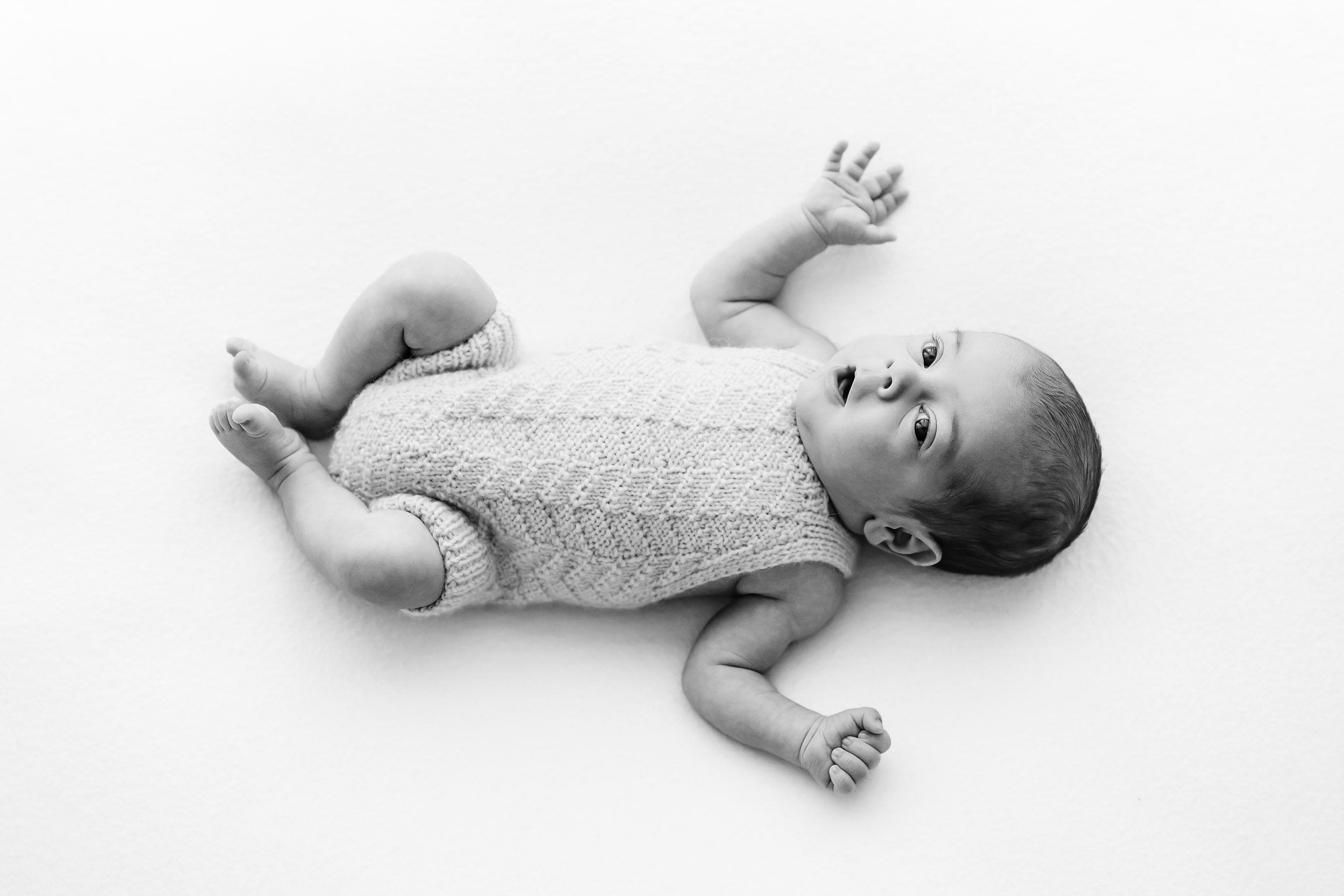 a black and white picture of a baby boy wearing a textured white knit romper laying on his back and holding his arms out as he looks up at the camera during a natural newborn photoshoot