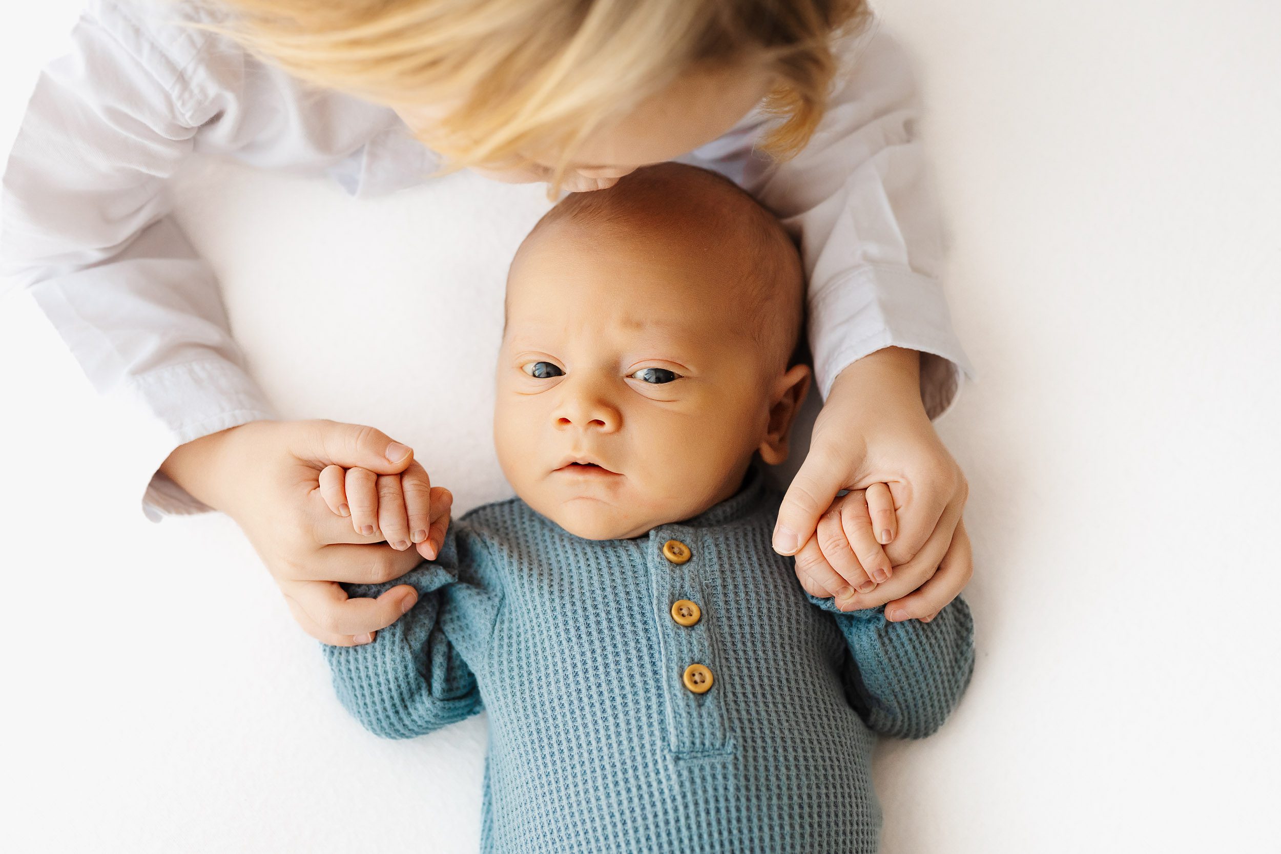 a baby boy wearing a long sleeve blue textured bodysuit laying on his back and holding onto his big brother's fingers as his brother gives him a kiss on the head during a natural newborn photoshoot
