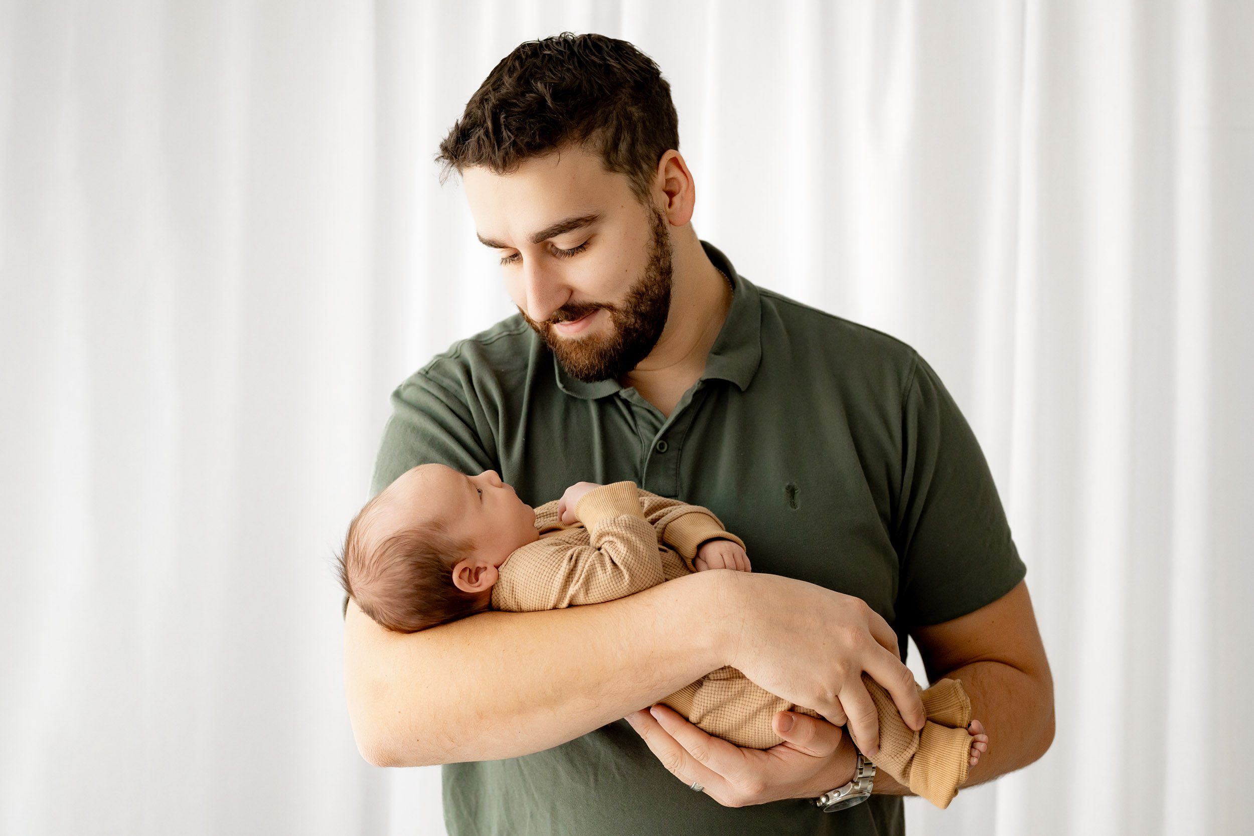a new dad cradling his baby boy in his arms and smiling down at him during a natural newborn photoshoot