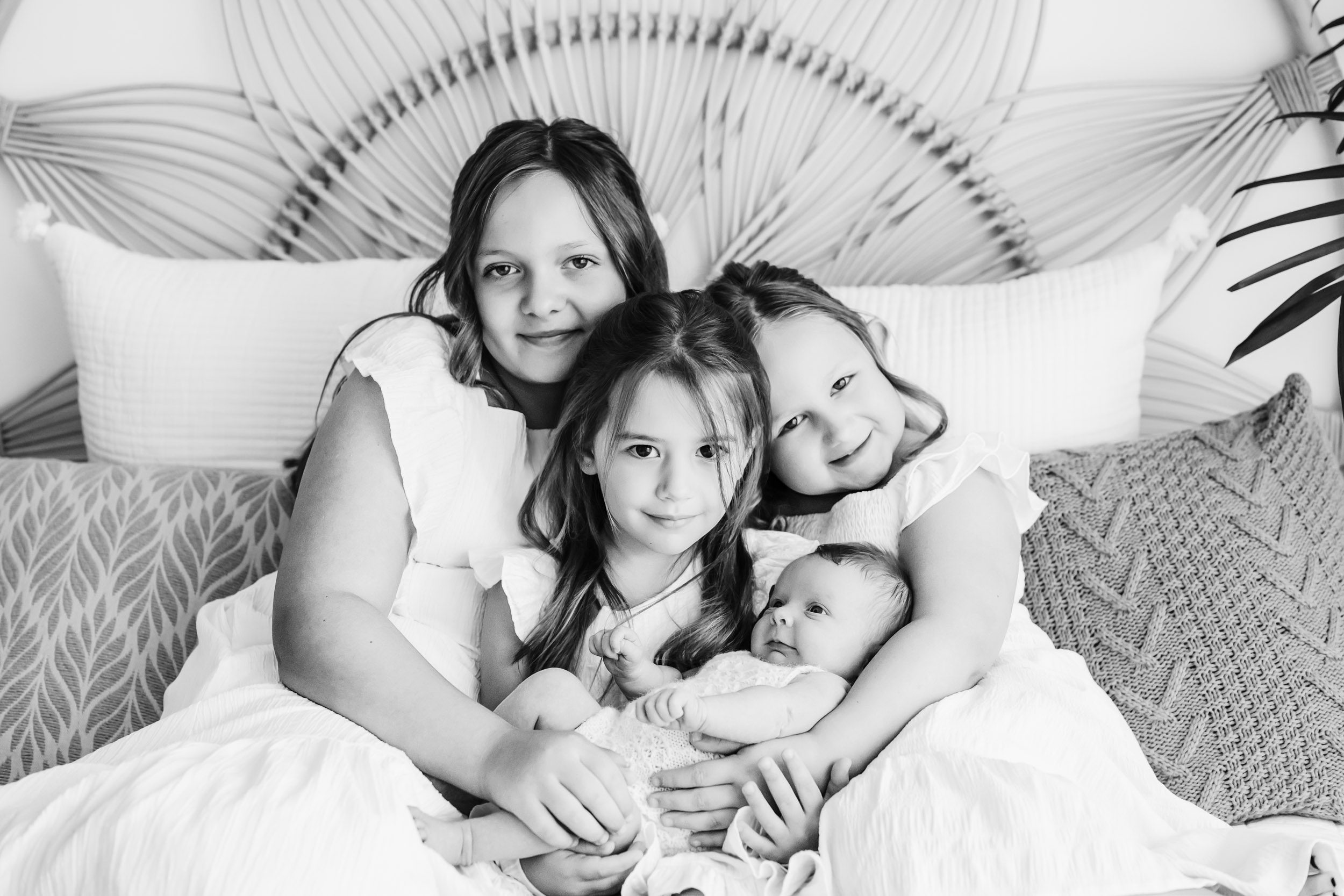 a black and white picture of three big sisters sitting together on a bed smiling at the camera with their baby sister cradled in their arms during a natural newborn photoshoot
