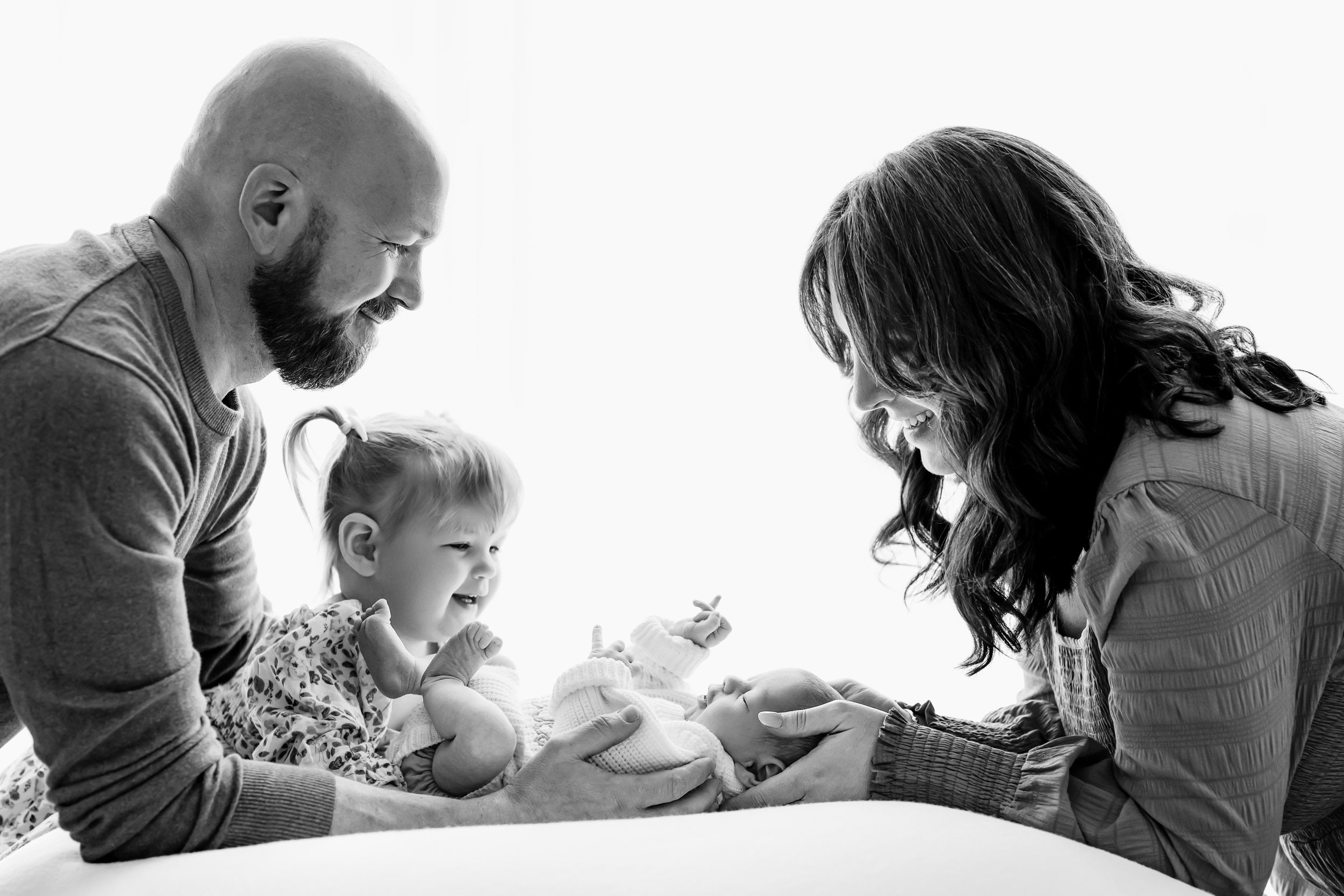a black and white backlit photo of a baby boy laying on a bean bag with his mom, dad, and big sister smiling down at him during a natural newborn photoshoot