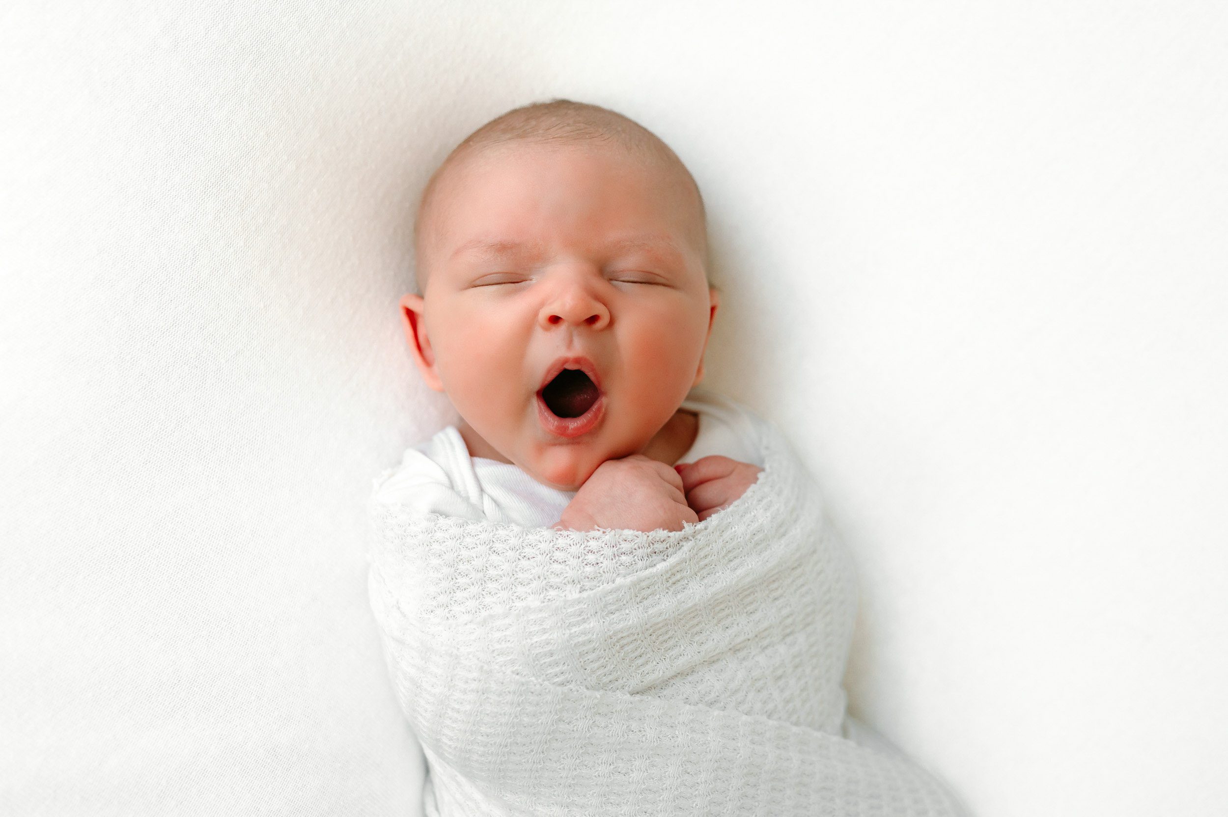 Yawning newborn baby boy in a white textured swaddle laying on a white backdrop during his natural newborn photos