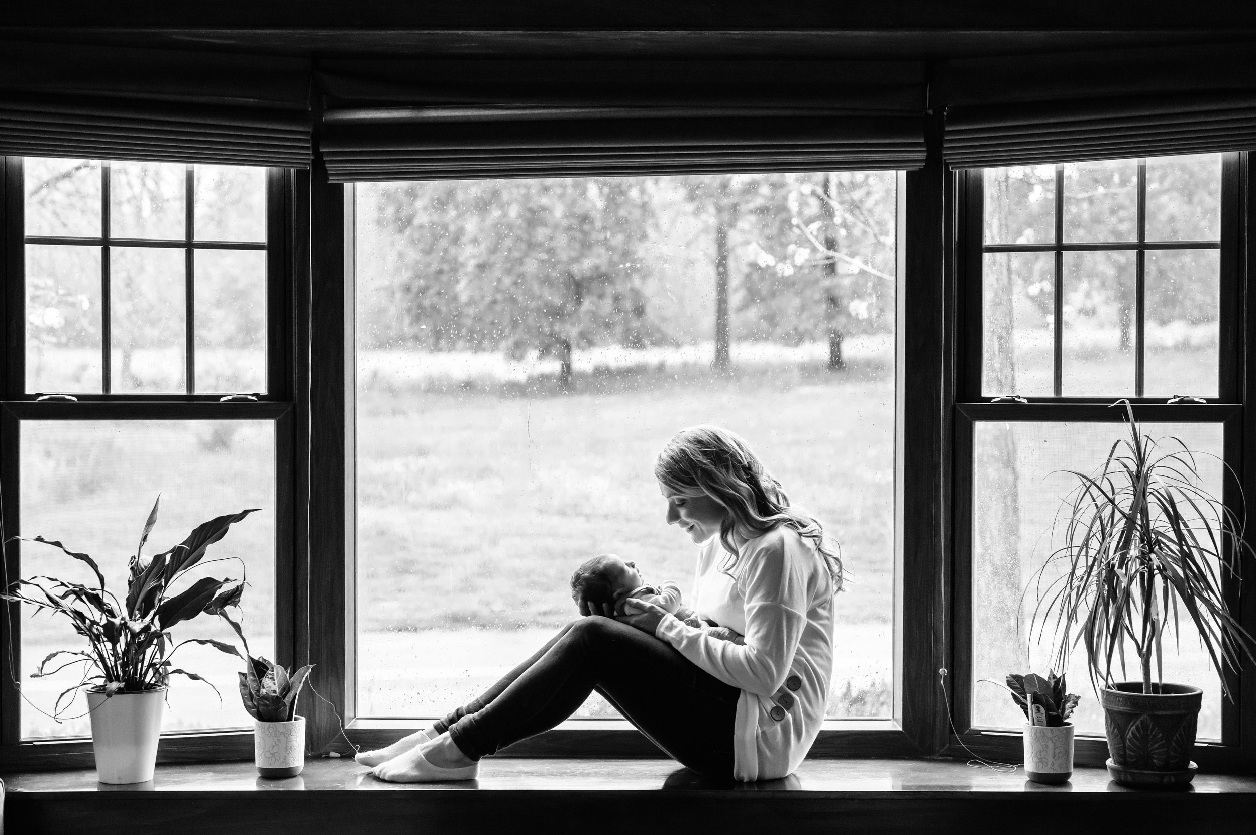 A black and white image of a new mother sitting in a bay window smiling at her newborn baby boy in her lap during a west chester home newborn session
