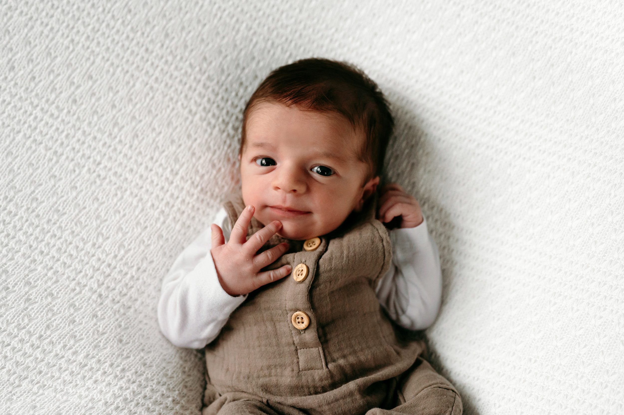 A newborn baby boy in a taupe jumper laying on a white backdrop and looking at the camera as he touches his lips with his finger during a west chester newborn photoshoot
