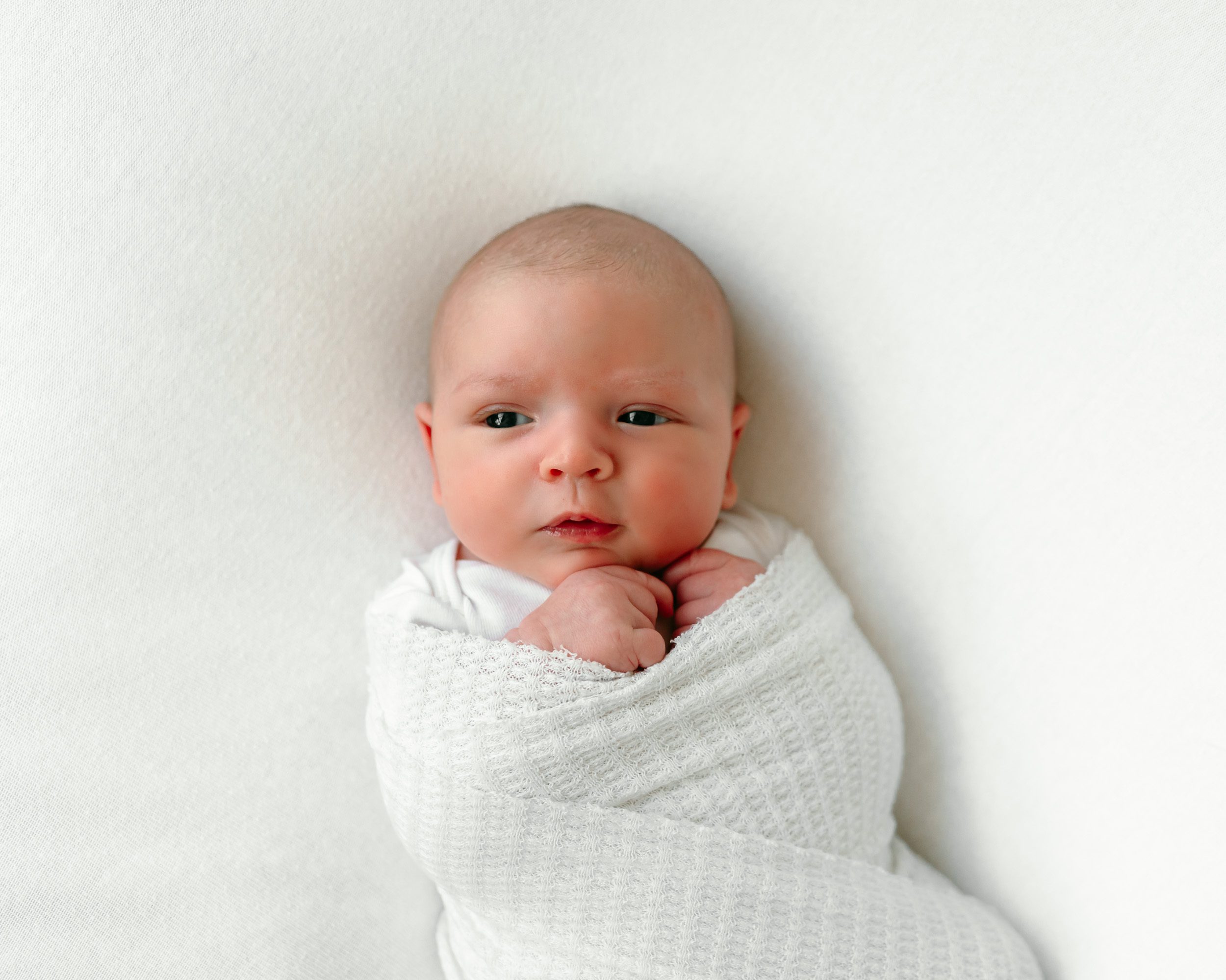 An older baby wrapped in a white textured swaddle laying on a white backdrop during a newborn photos session