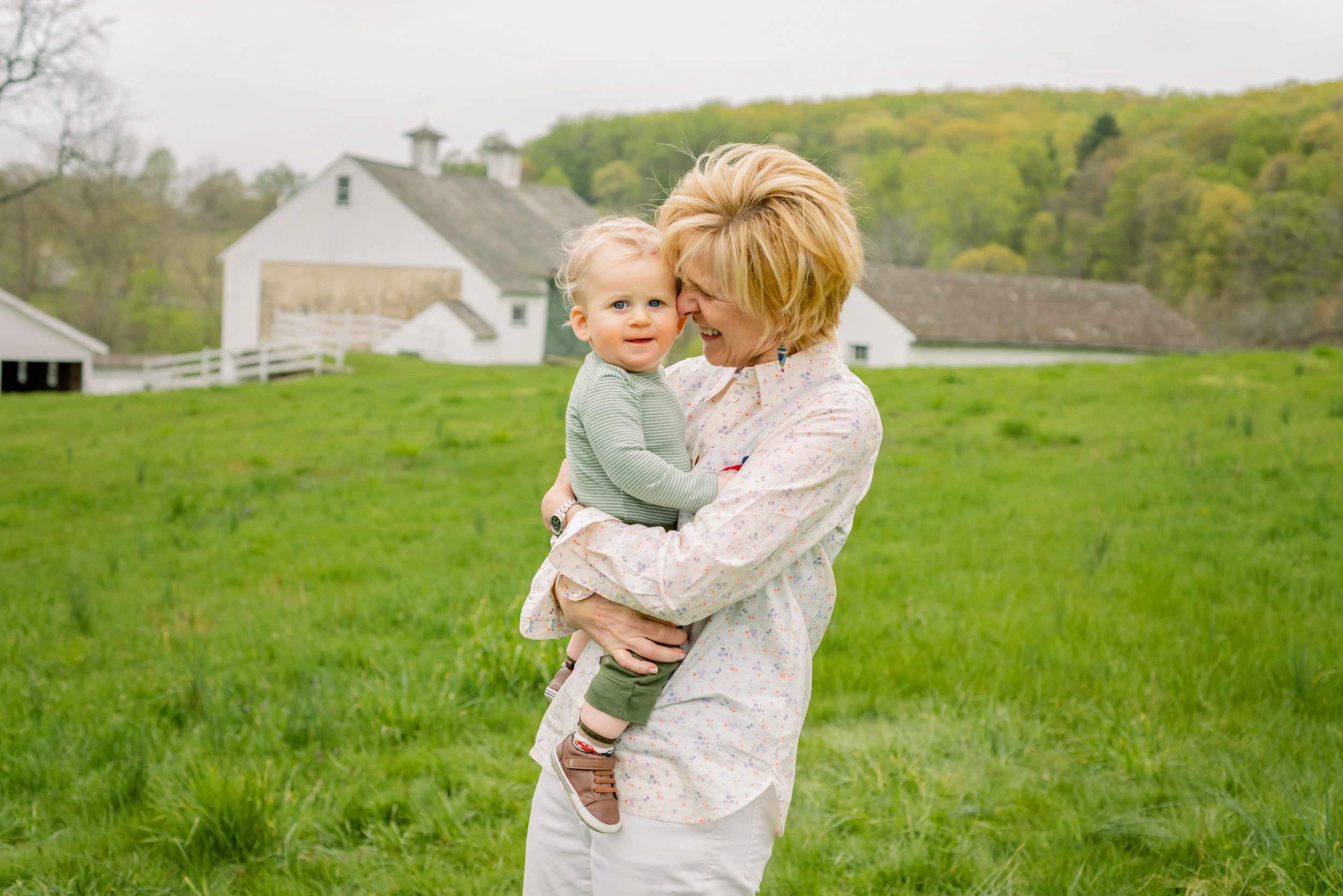 A grandmother snuggling her grandson with white and green barns in the background during an extended family photo session
