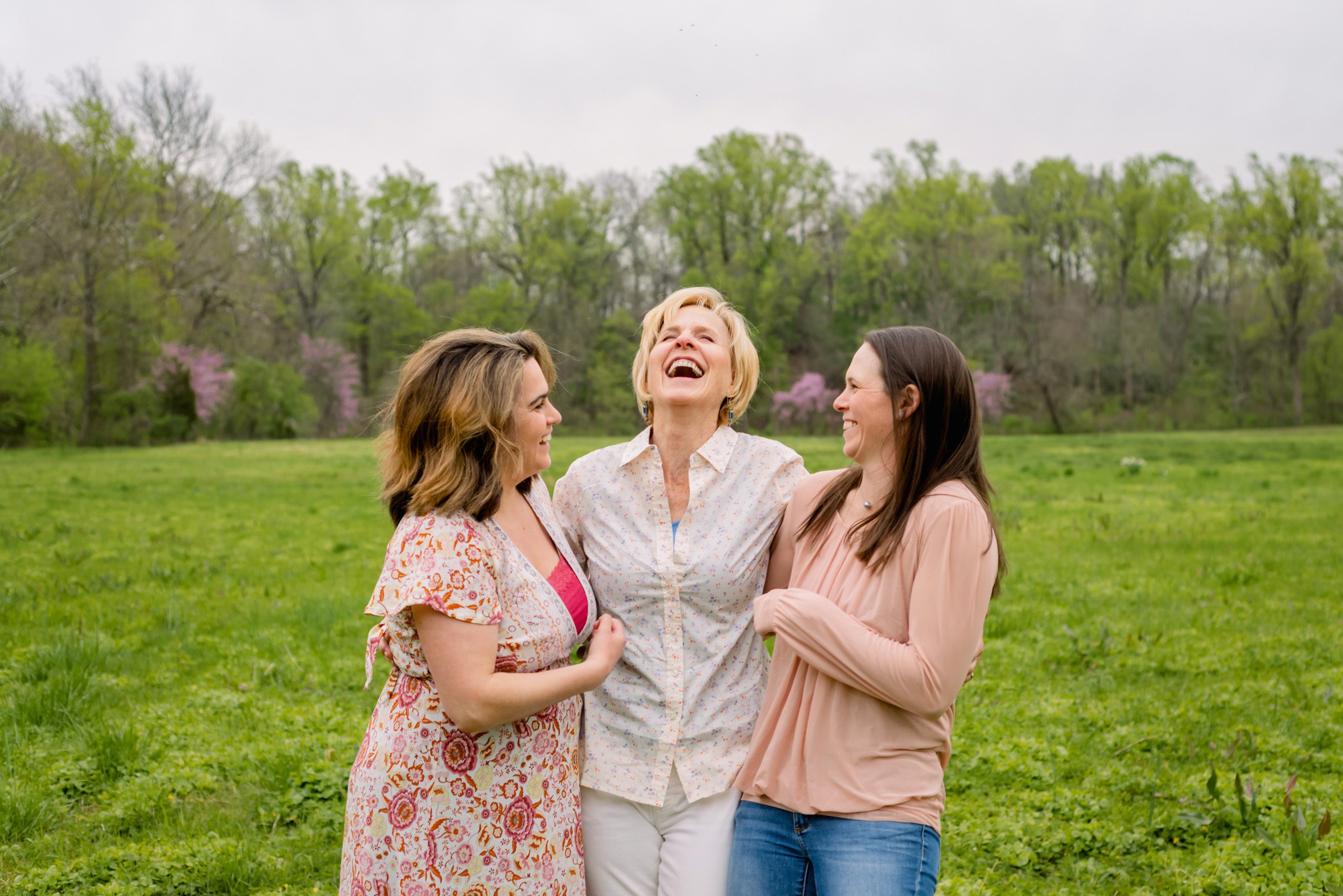 A mom and her two adult daughters laughing in a grassy field with redbud trees blooming in the background during an extended family photoshoot