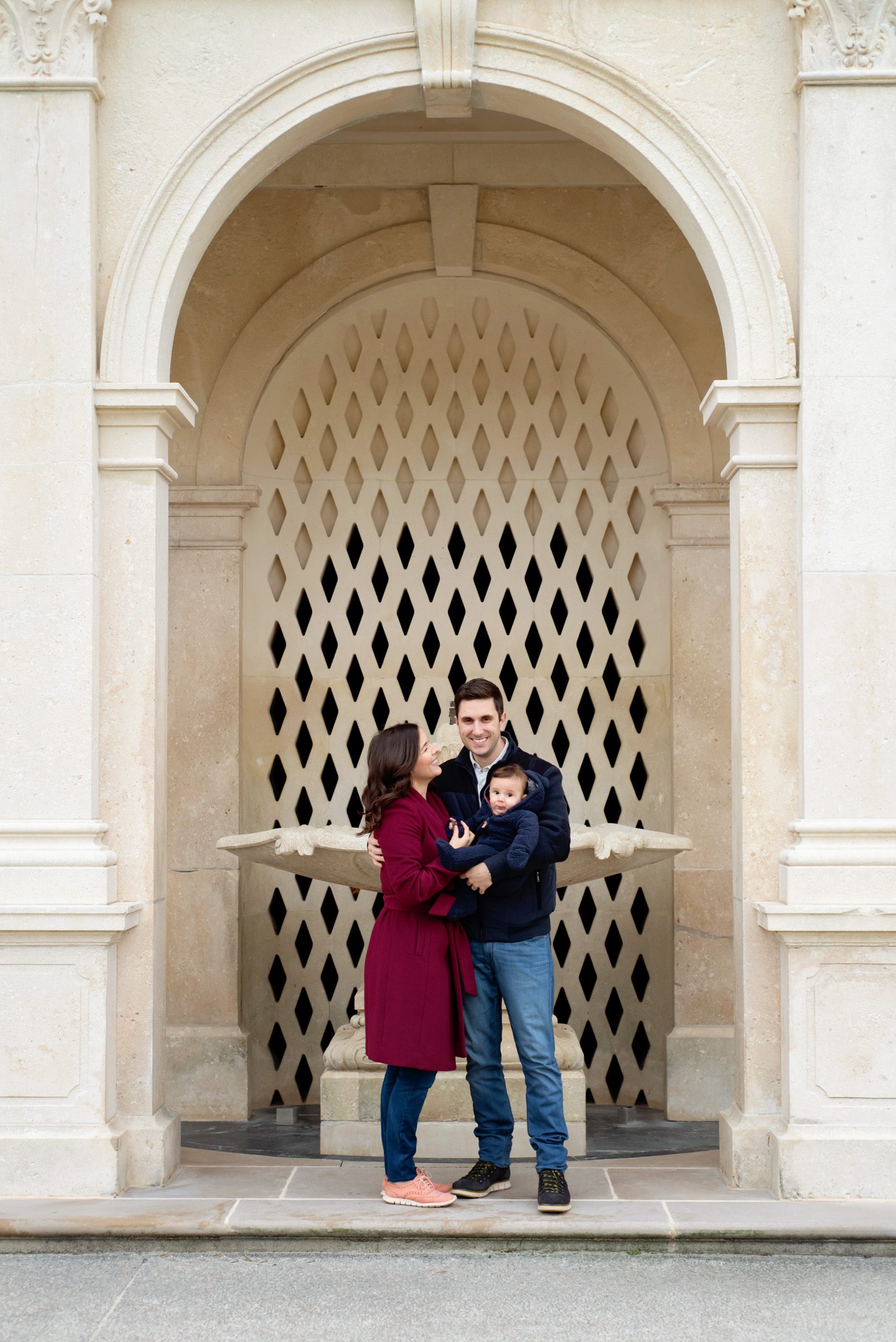 Parents standing in a white marble archway holding their young son and smiling at each other during a family photo session
