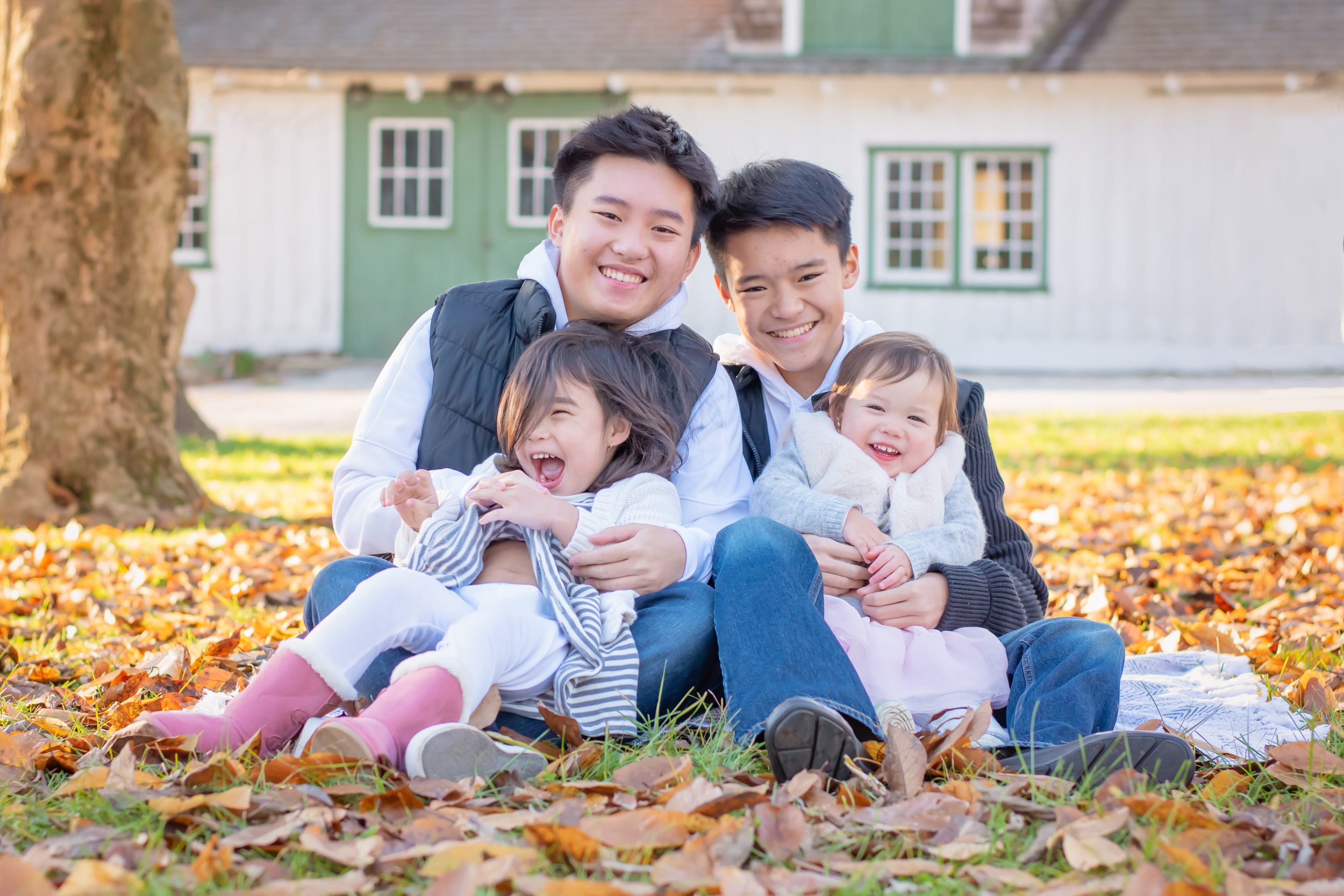 Cousins sitting in a field of leaves snuggling and laughing with a green and white barn in the background during an extended family photo session