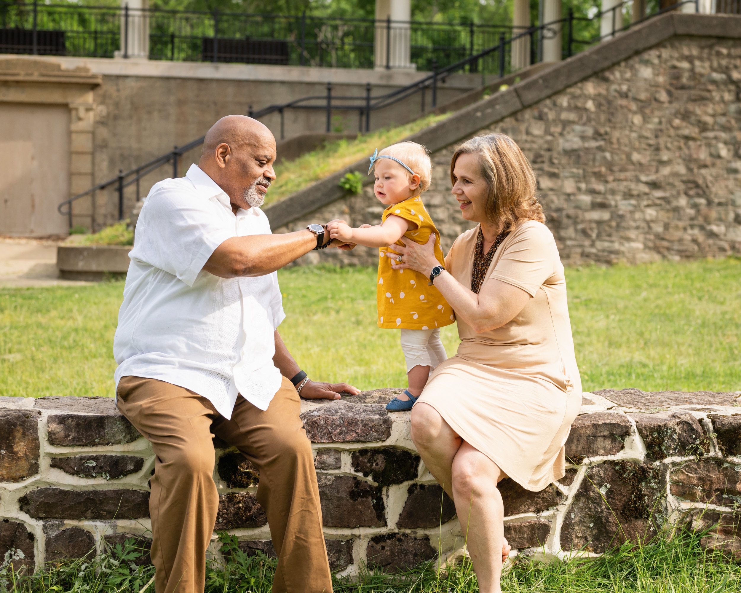 Grandparents sitting on a stone wall with their granddaughter as she examines her grandfather's watch during an extended family photo session