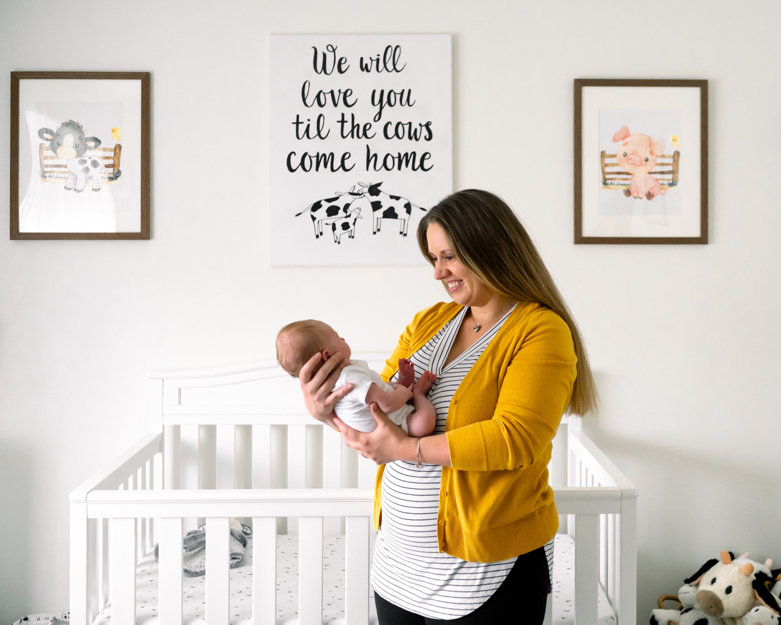 A mom smiling down at her newborn son in her arms while standing in front of the crib in his nursery during an in home newborn photo session