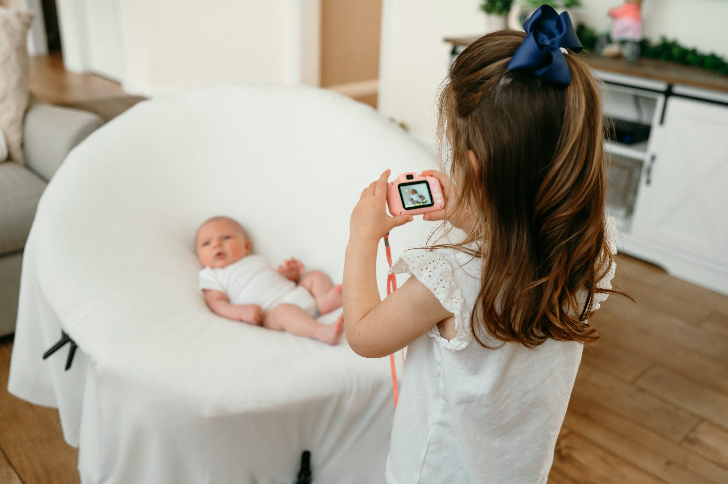 A newborn baby boy in a white onesie laying in a newborn photographer's photo chair while his big sister takes a picture of him with her toy camera during a natural newborn photo session