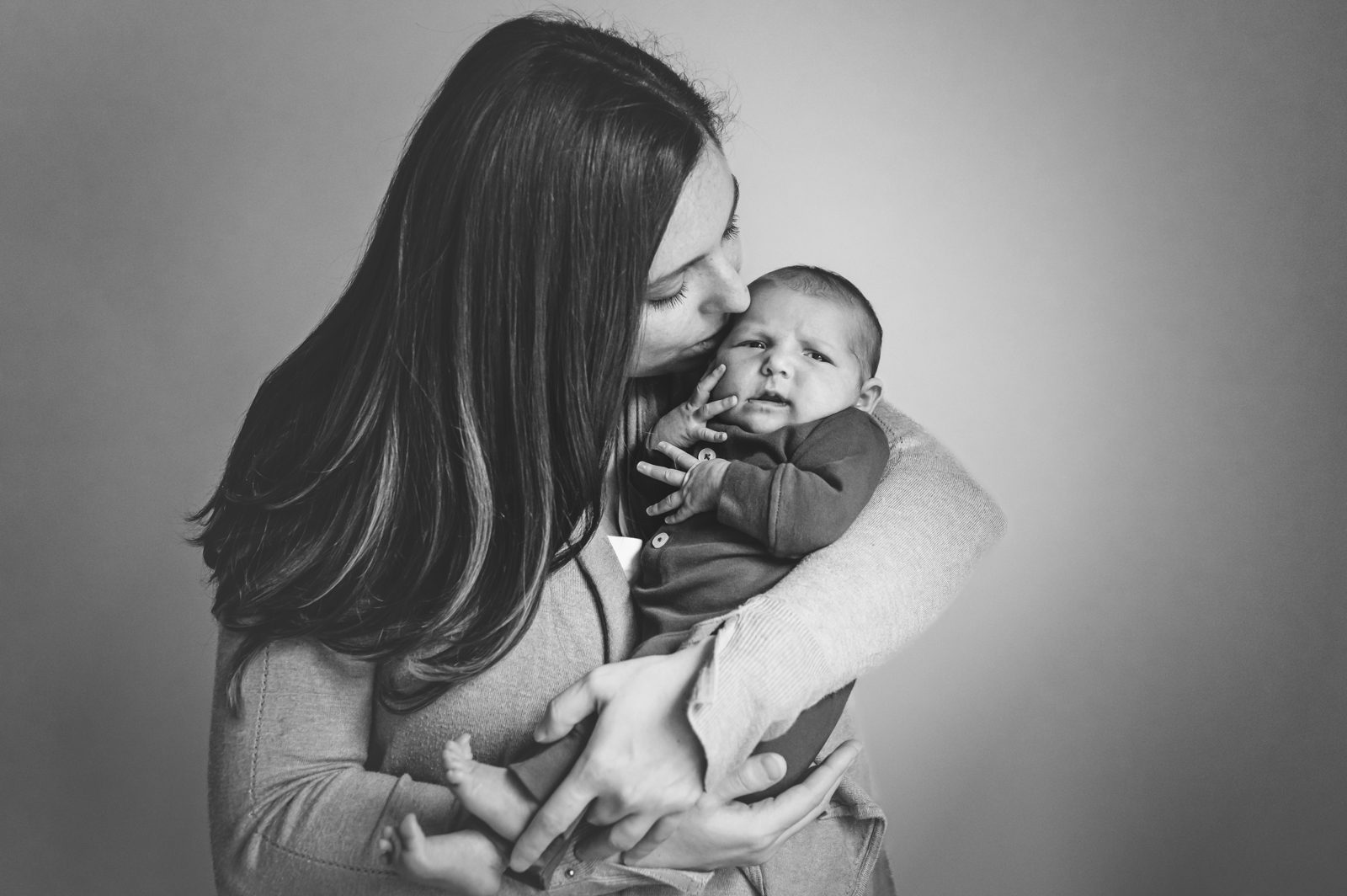 Black and white image of a mother cradling her newborn baby girl and kissing her on the temple during a natural newborn photoshoot
