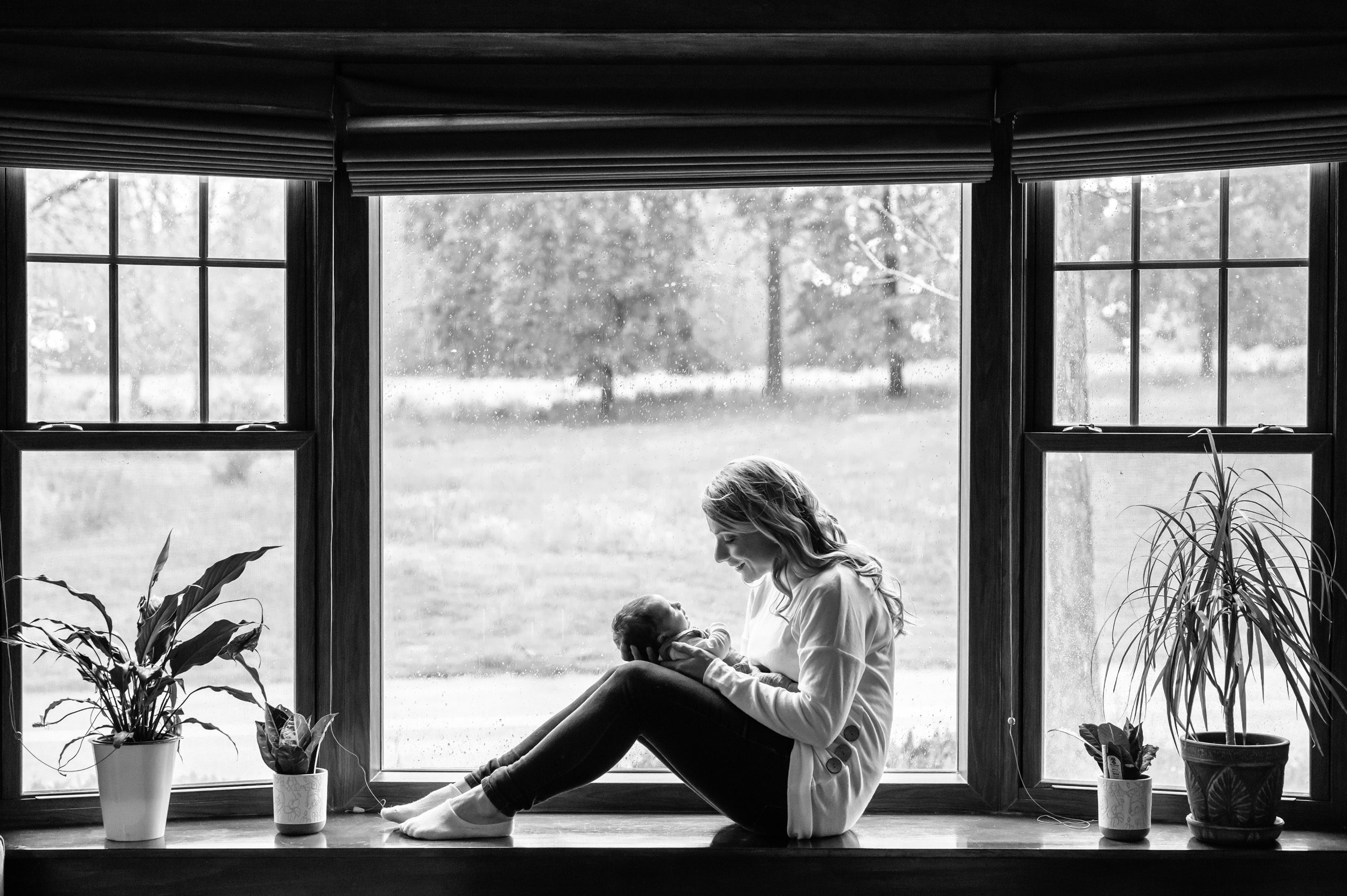 Black and white image of a mom sitting in a bay window and smiling down at her newborn baby boy during a natural newborn photoshoot