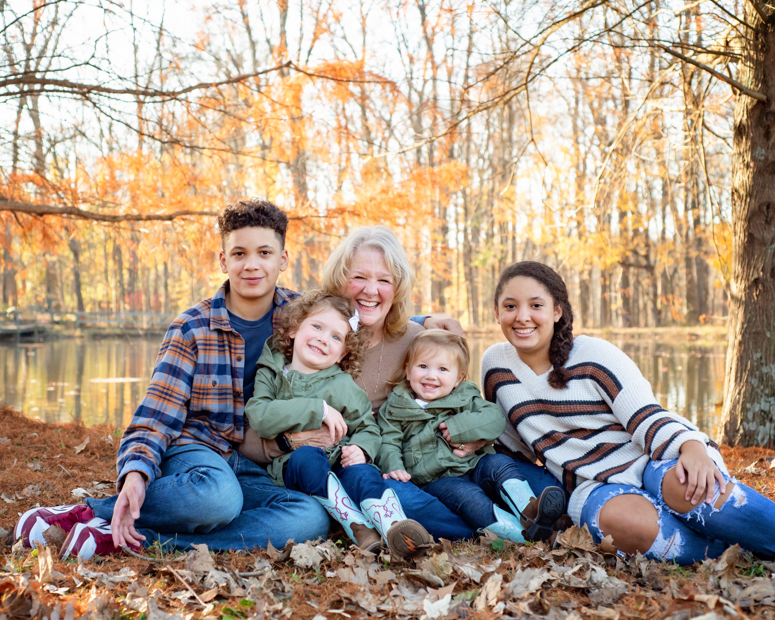 A grandmother sitting in the leaves in front of a pond and snuggling with her four grandkids during an extended family photoshoot