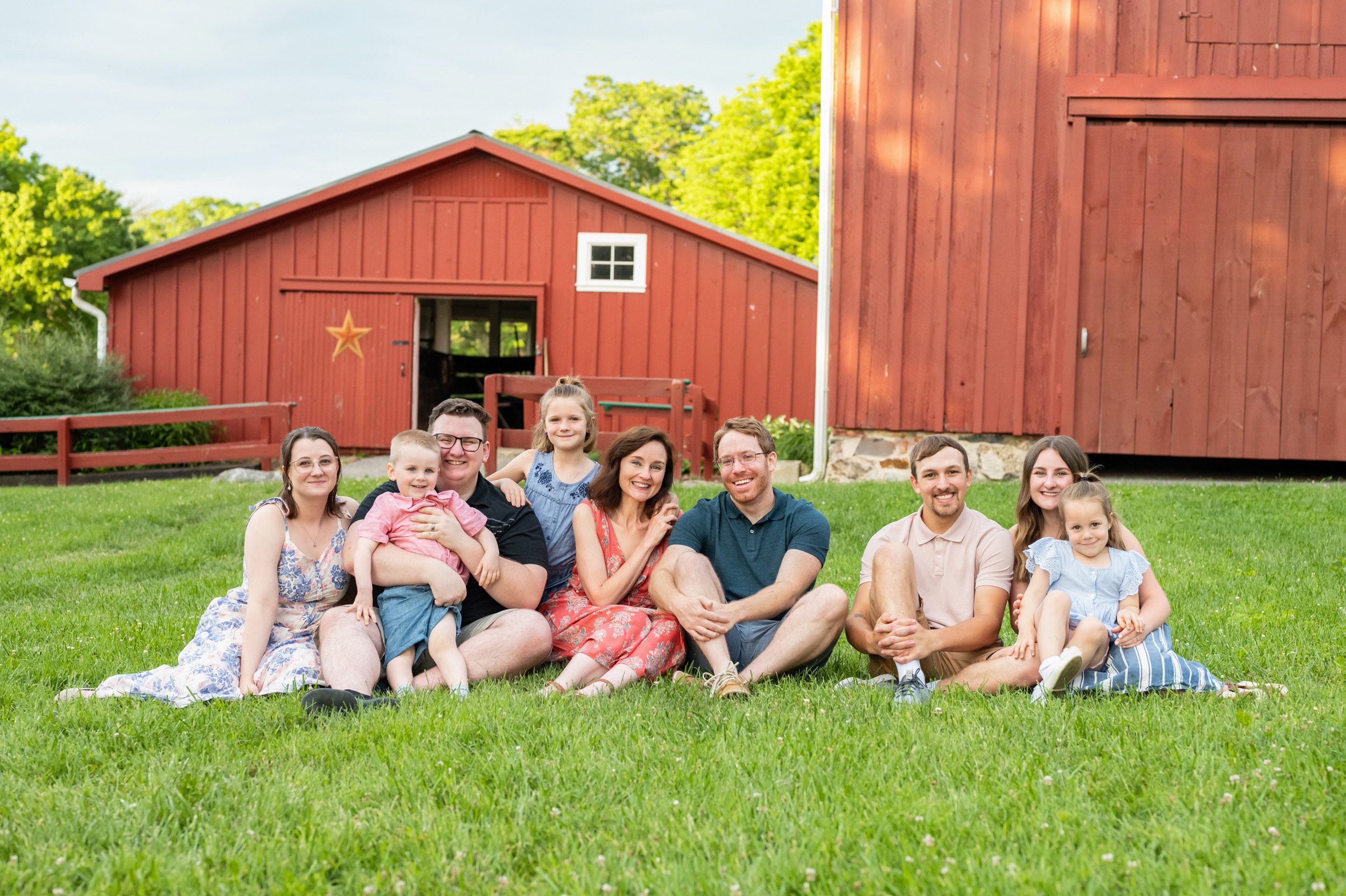 An extended family with grandparents, their kids and spouses, and their grandkids sitting in the grass in front of a red barn during an extended family photoshoot
