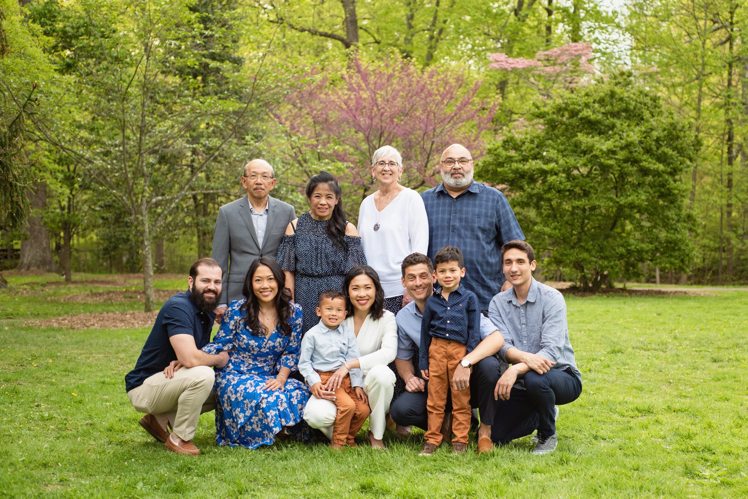 An extended family with both sets of grandparents standing behind their kids and grandkids in a grassy field with redbud trees blooming in the background during an extended family photo session