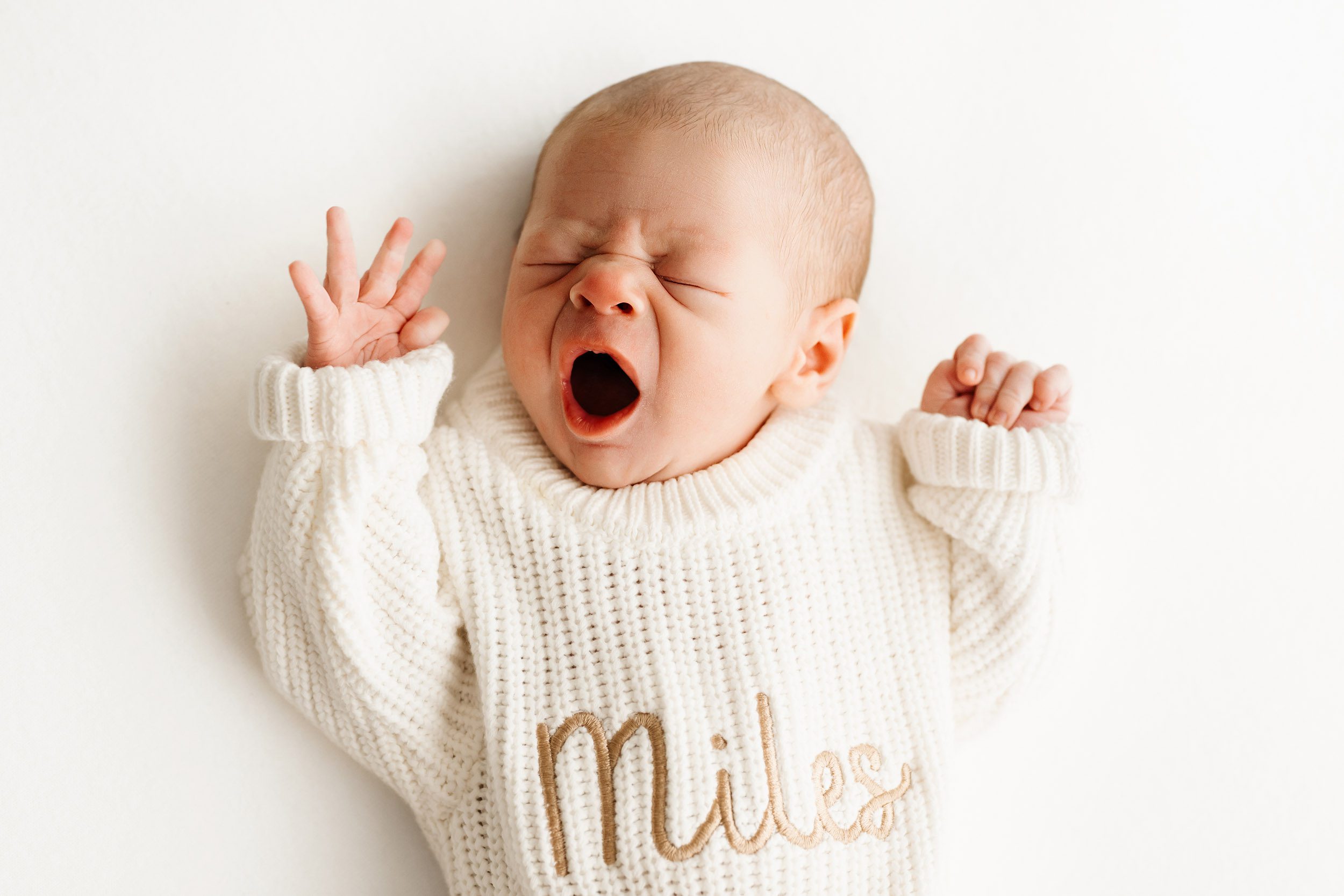 a baby boy wearing a knit sweater with Miles written on it laying on a white backdrop and yawning during a newborn photography session