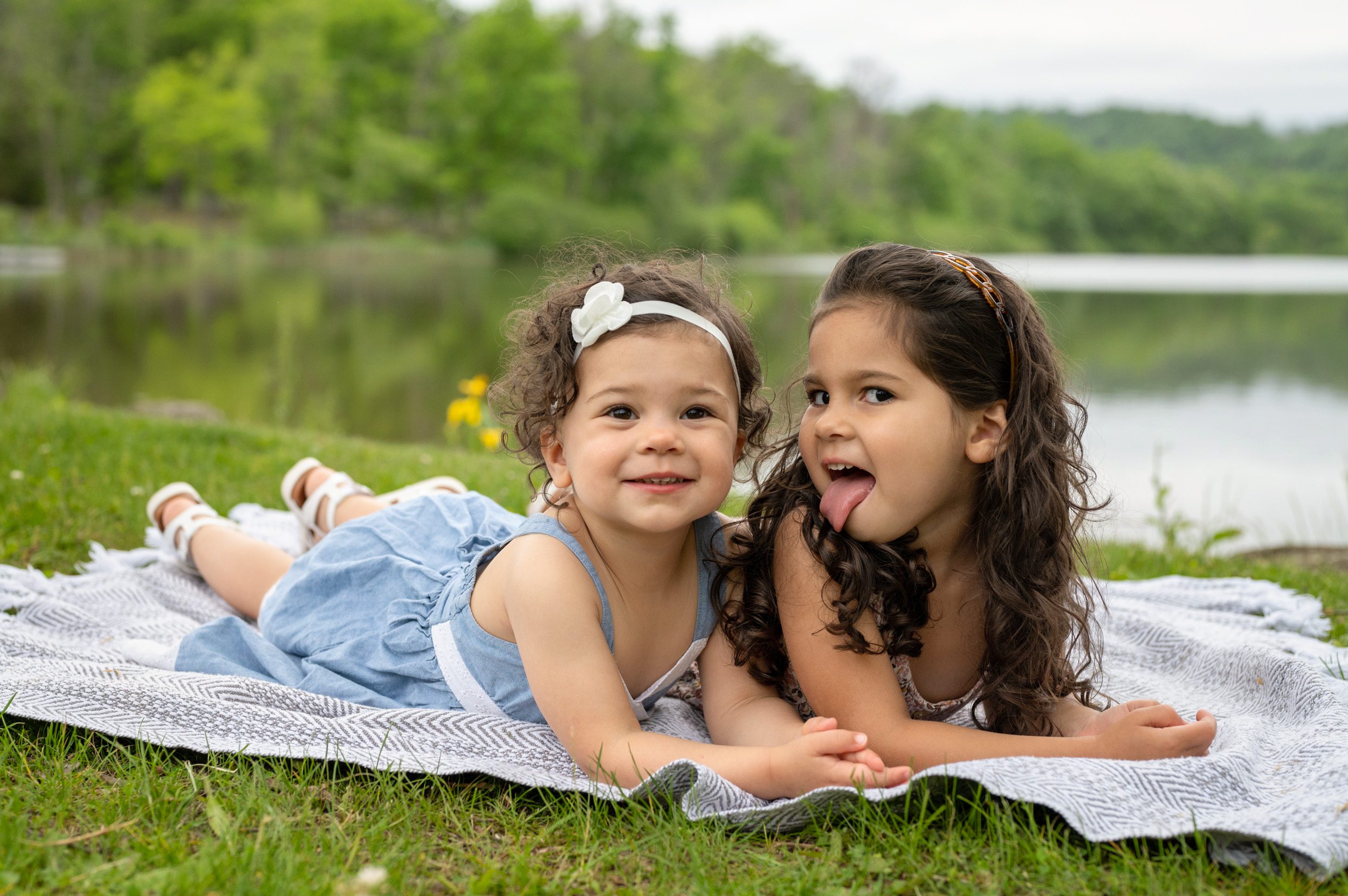 Two sisters laying next to each other on a blanket in front of a lake while one sister sticks out her tongue and the other sister smiles during a family photoshoot