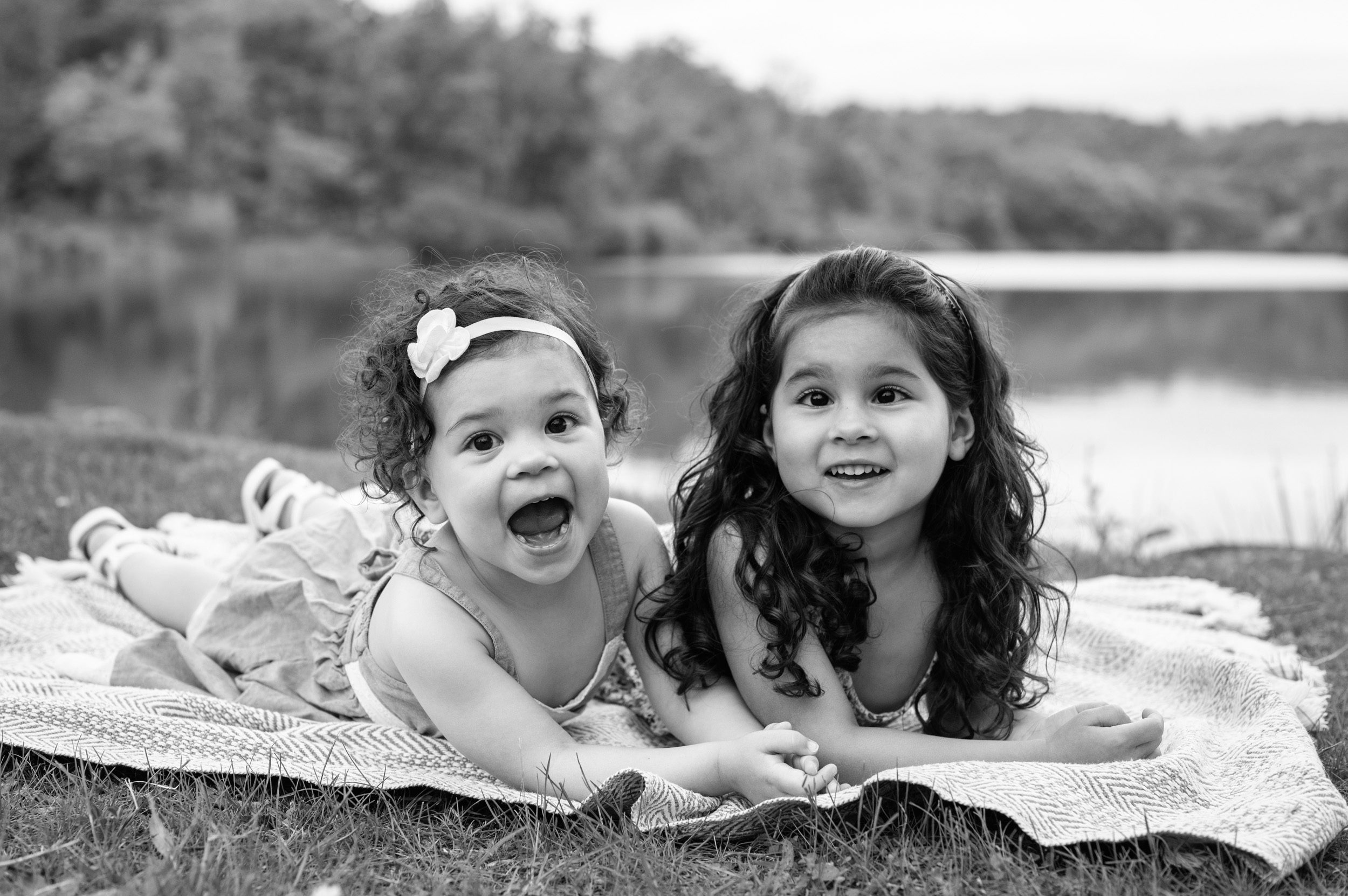 a black and white picture of two young sisters laying next to each other on a blanket and making silly faces at the camera during their family photos