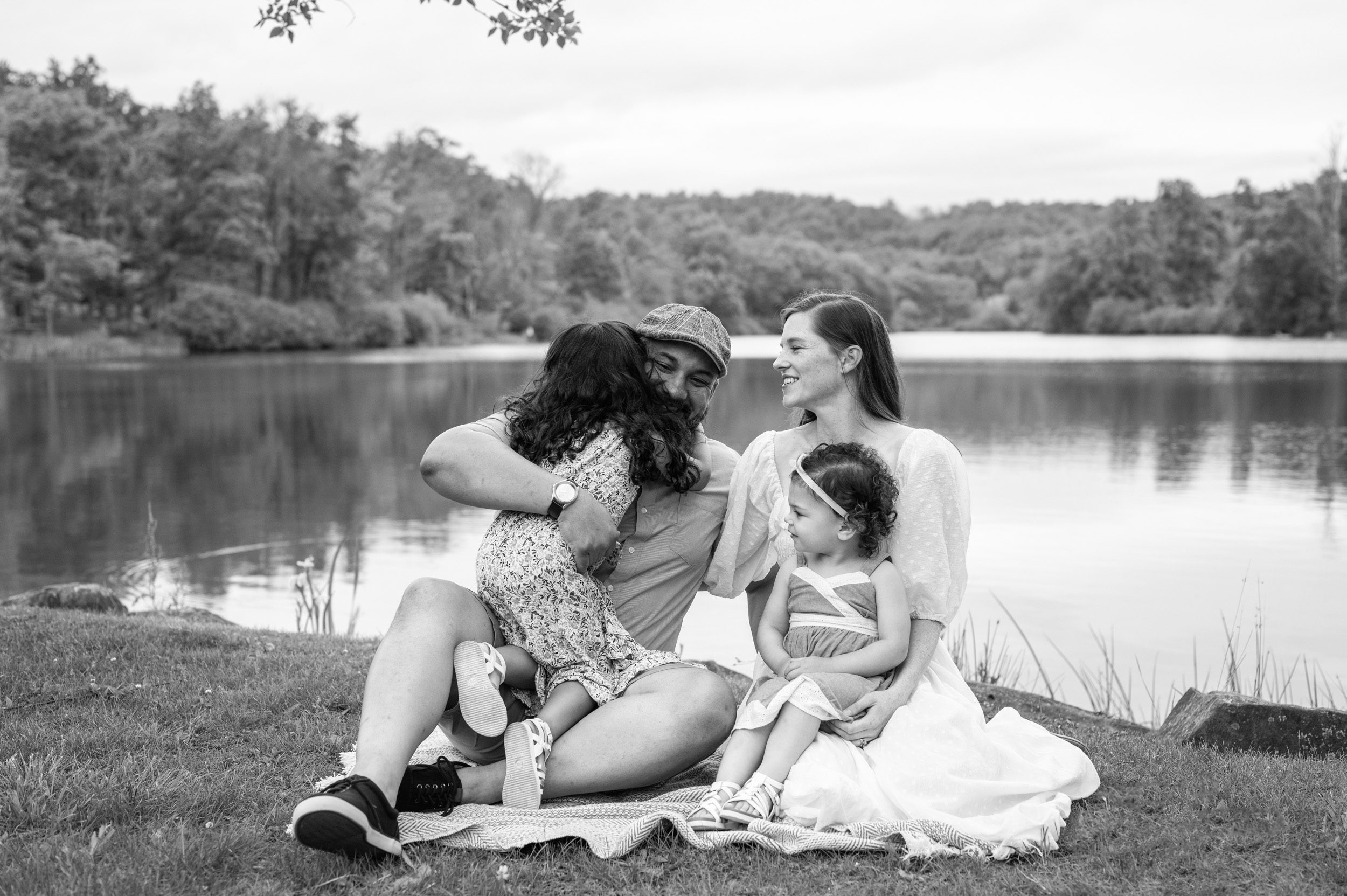Two young girls sitting on a blanket in front of a lake and hugging their parents during a pottstown family photoshoot 