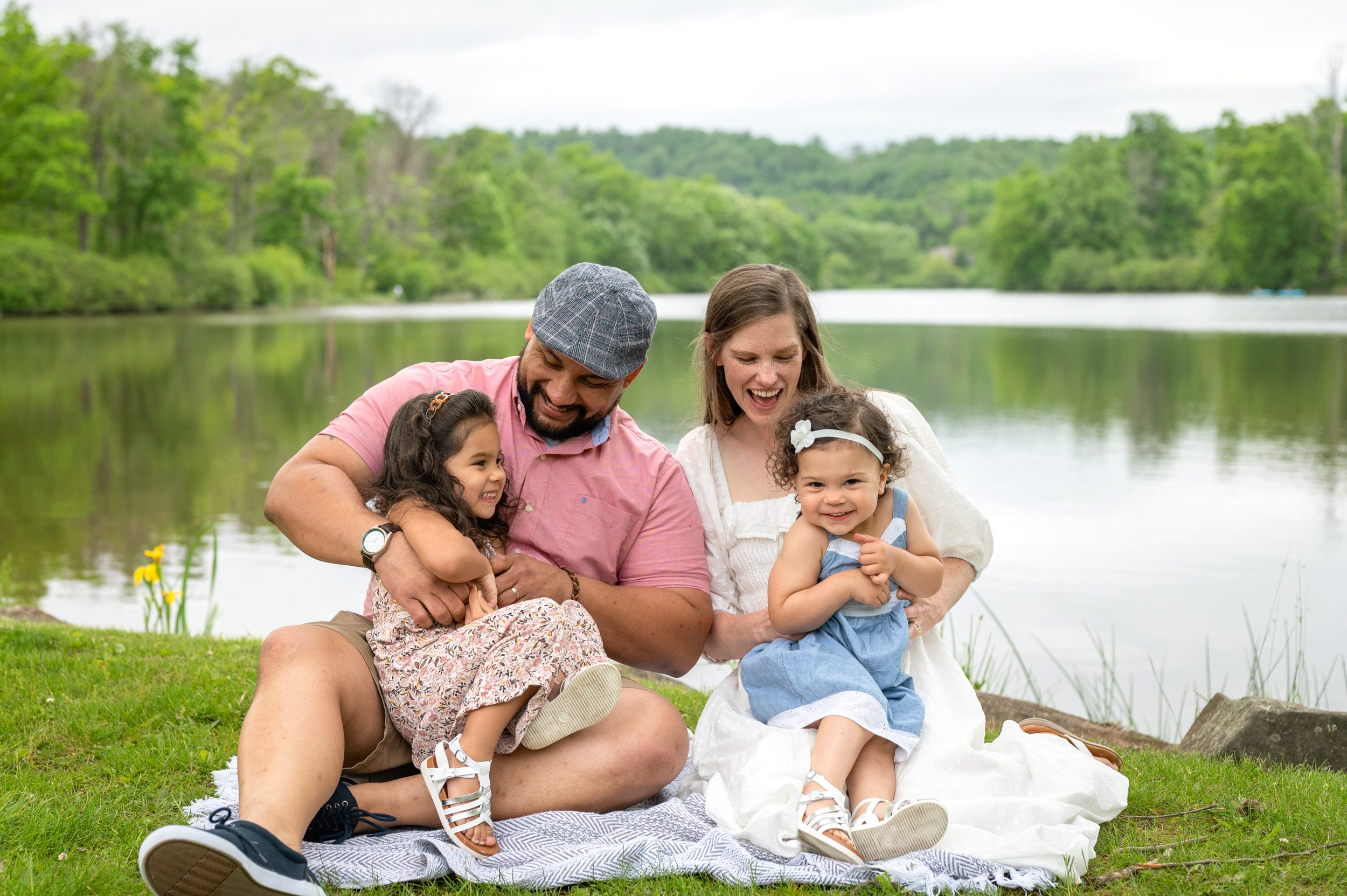 Two parents sitting on a blanket in front of a lake tickling their young daughters on their lap during a family photo session