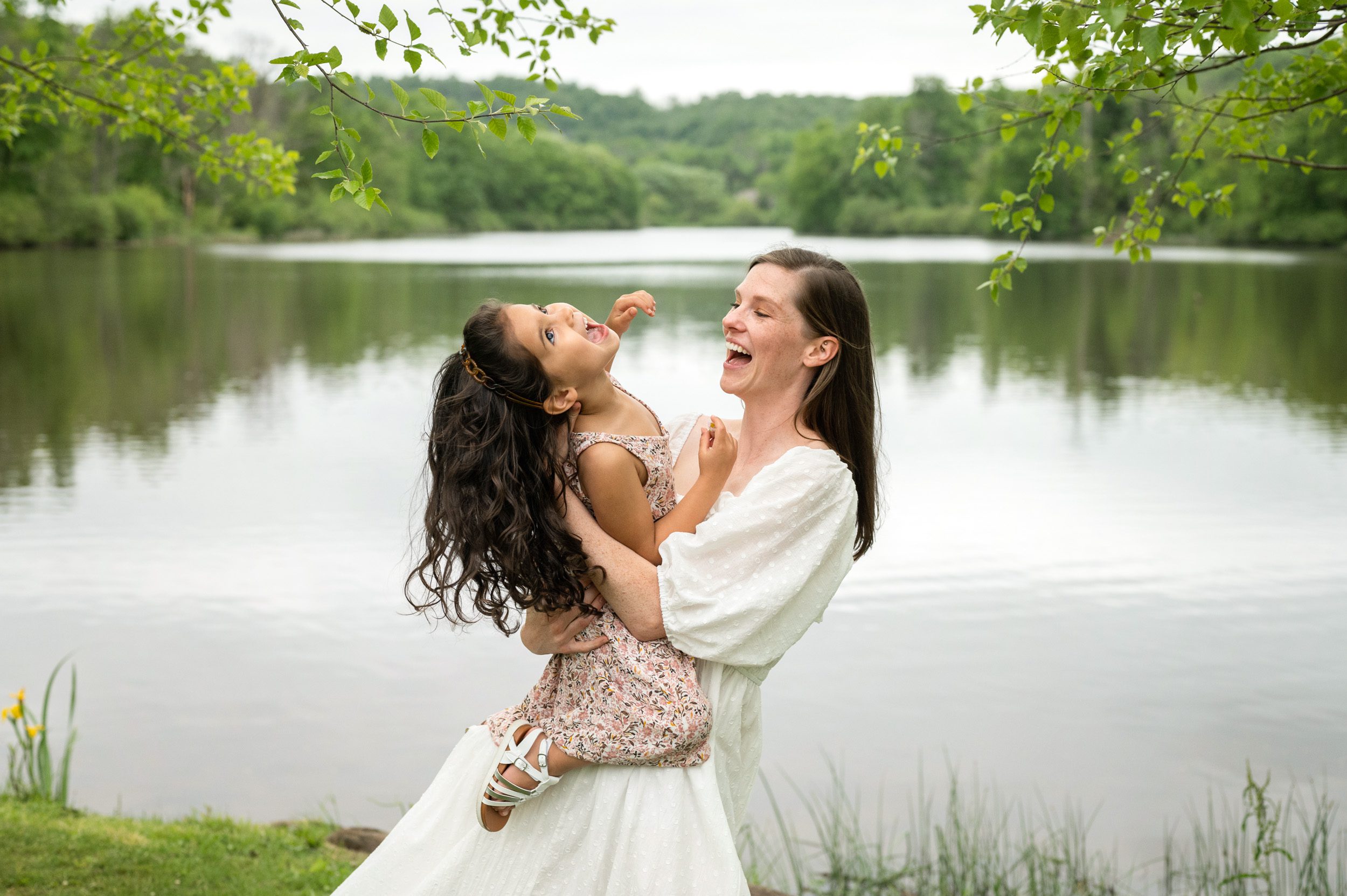 A mom standing in front of a lake and dipping her young daughter back as they both laugh during a family photoshoot
