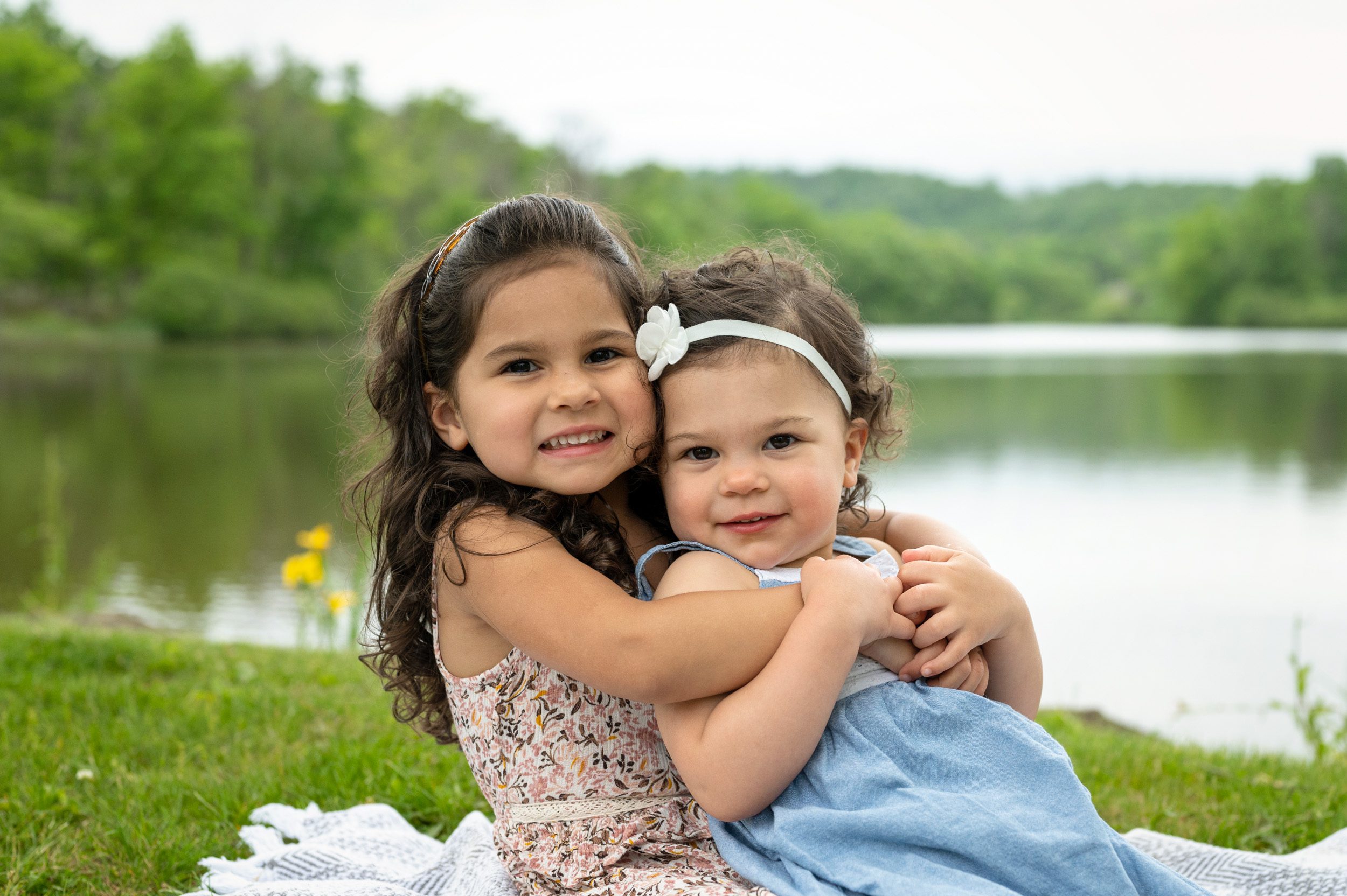 Two sisters sitting on a blanket and hugging in front of a lake during their family photos