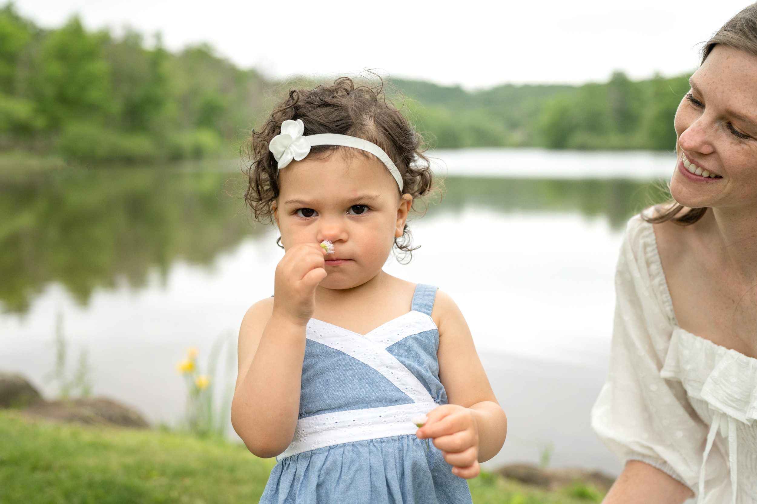 A little girl smelling a flower with a serious expression while her mom smiles at her during a family photoshoot