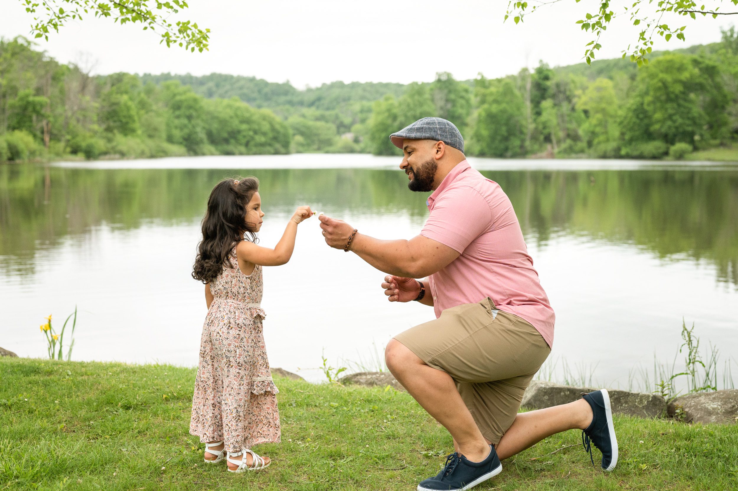 A little girl and her daddy standing in front of a lake and touching the flowers in their hands together during a pottstown family photo session