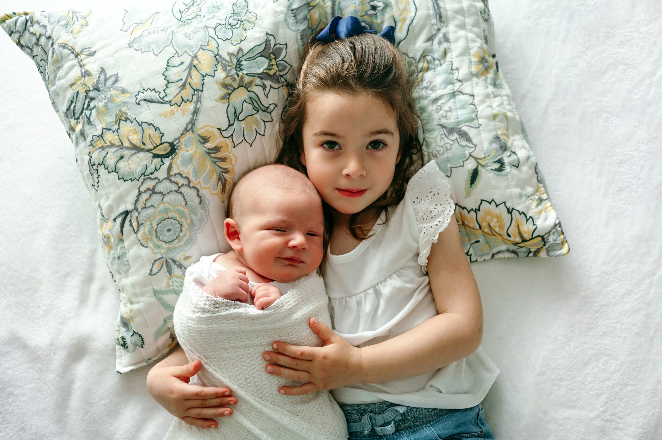 An older sister and her newborn baby brother laying on a bed and looking up at the camera during a home newborn photo session