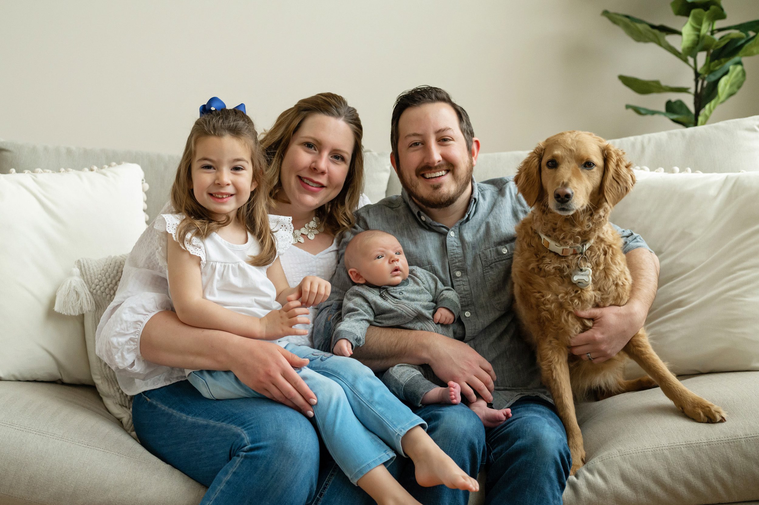 family sitting on their couch with their older daughter, pet dog, and newborn baby boy during a home newborn photo session