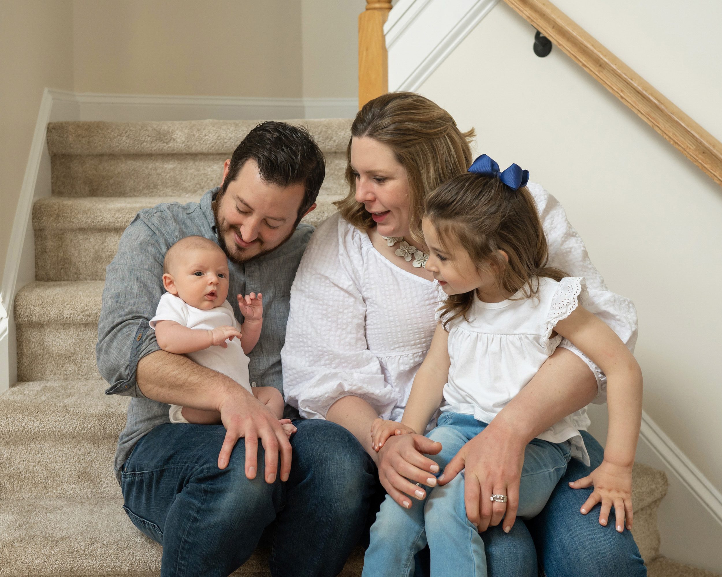 A new family of four sitting on their steps and smiling at their newborn baby boy during a gilbertsville home newborn photo session