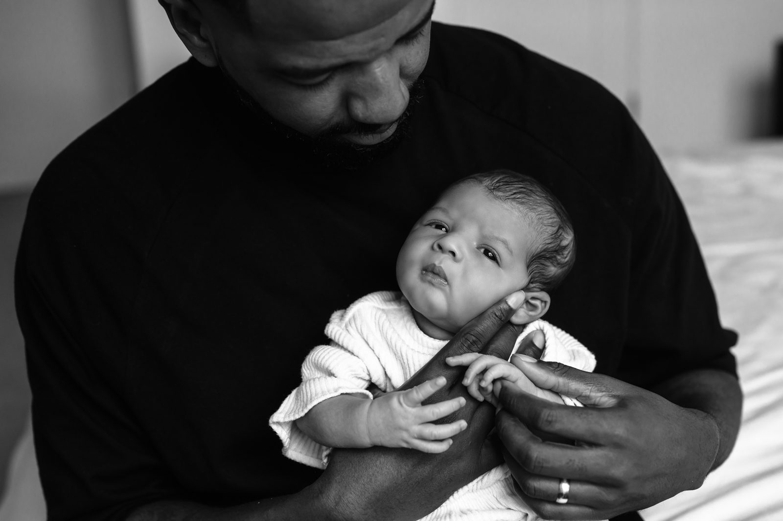 a black and white picture of a dad holding his newborn baby girl with her back against his chest as she grabs onto his finger during an in home lifestyle newborn session